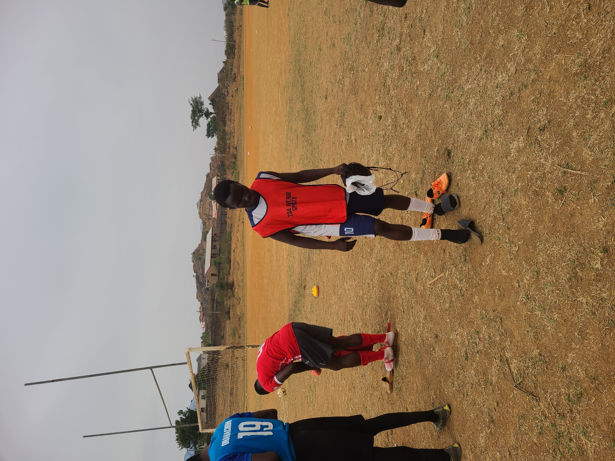 The scene is an outdoor field in Barikin Jirga Summa, Nigeria. Three individuals are visible. One person wears a red practice vest with the text "234 AVENUE SPORTS" and the number "10" on their shorts. The person appears to be holding a pair of headphones and some white athletic tape. Another person is bending over in a red jersey and black shorts. A third individual is wearing a blue jersey with the number "19" visible. The field appears to be dry and grassy, with a goalpost visible in the background. In the distance, there are buildings and hills.