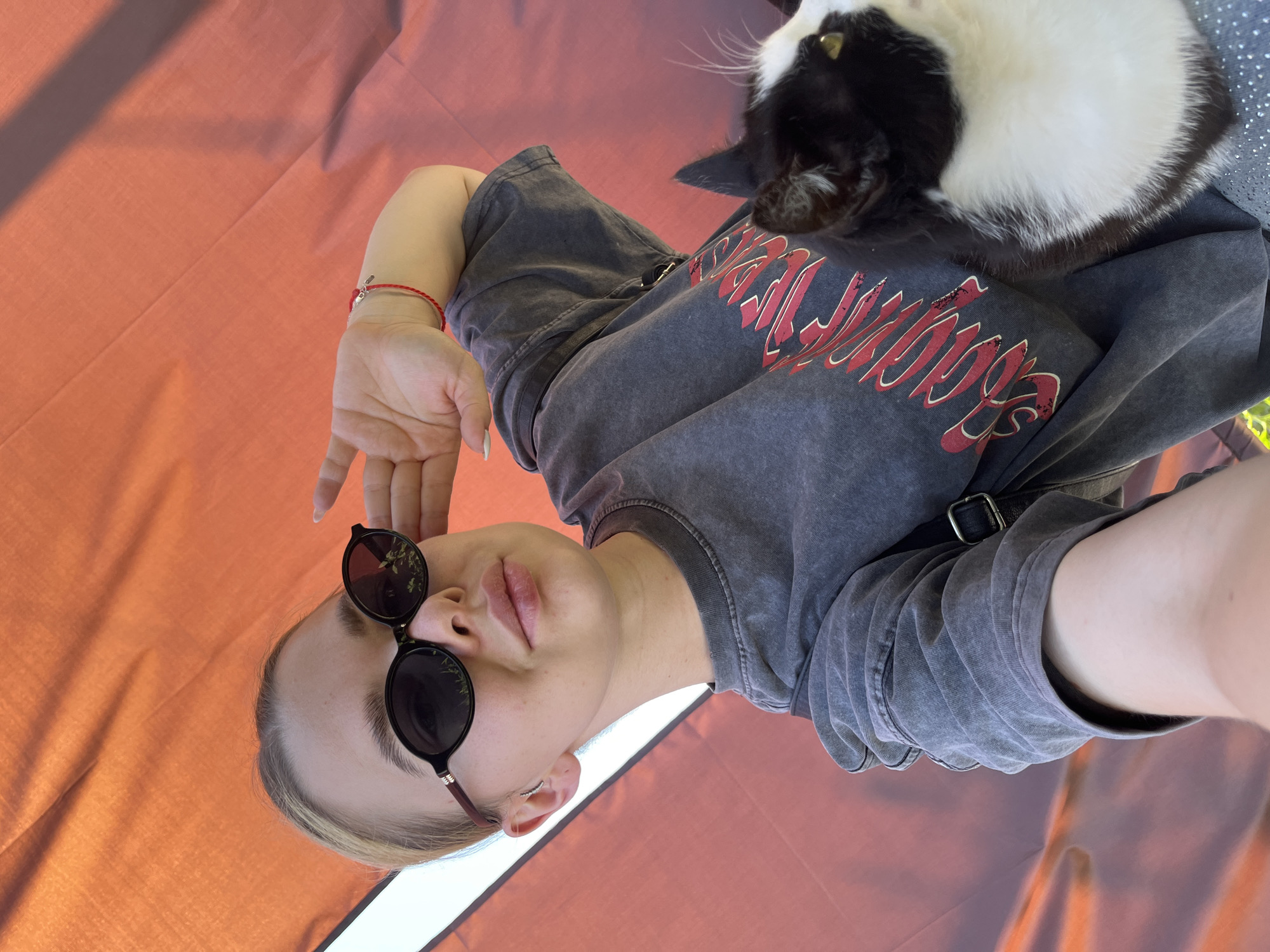 A young woman with blonde hair pulled back, wearing sunglasses and a dark gray t-shirt with a red graphic design, holds a black and white cat while taking a selfie outdoors. She is touching her forehead with her right hand, and a red bracelet is visible on her wrist. The background is an orange-red fabric, possibly an awning or umbrella, with some dappled sunlight. The scene appears to be in a casual outdoor setting. There is no specific information in the image to suggest the location is Tikhoretsk, Russia.