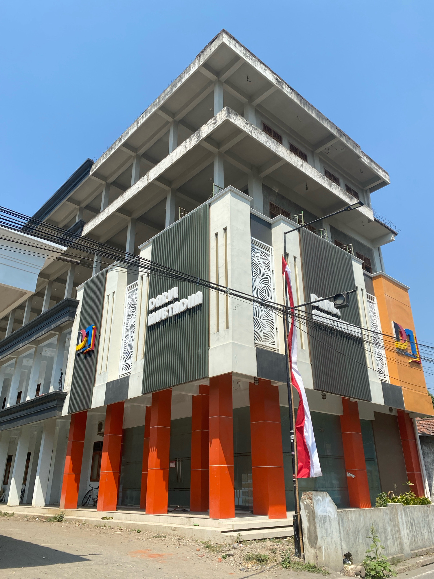 The image shows the front of a multi-story building with a bright orange and dark green facade. The building has a modern design with decorative white panels and vertical orange columns on the lower level. A prominent sign on the building reads "DARUL MUSTAQIM" in white, raised letters. A logo, appearing to be "DI," is also visible on the left side of the building. A red and white Indonesian flag is mounted on a pole in front of the building. The building appears to be located in an urban environment, with electrical wires visible overhead and a paved street in the foreground. The sky is clear and blue. The building's upper floors are unfinished, with exposed concrete and structural beams, suggesting it may be under construction or renovation. A bicycle is parked near the entrance on the left side. The overall scene suggests a commercial or institutional building in Kediri City, Indonesia.