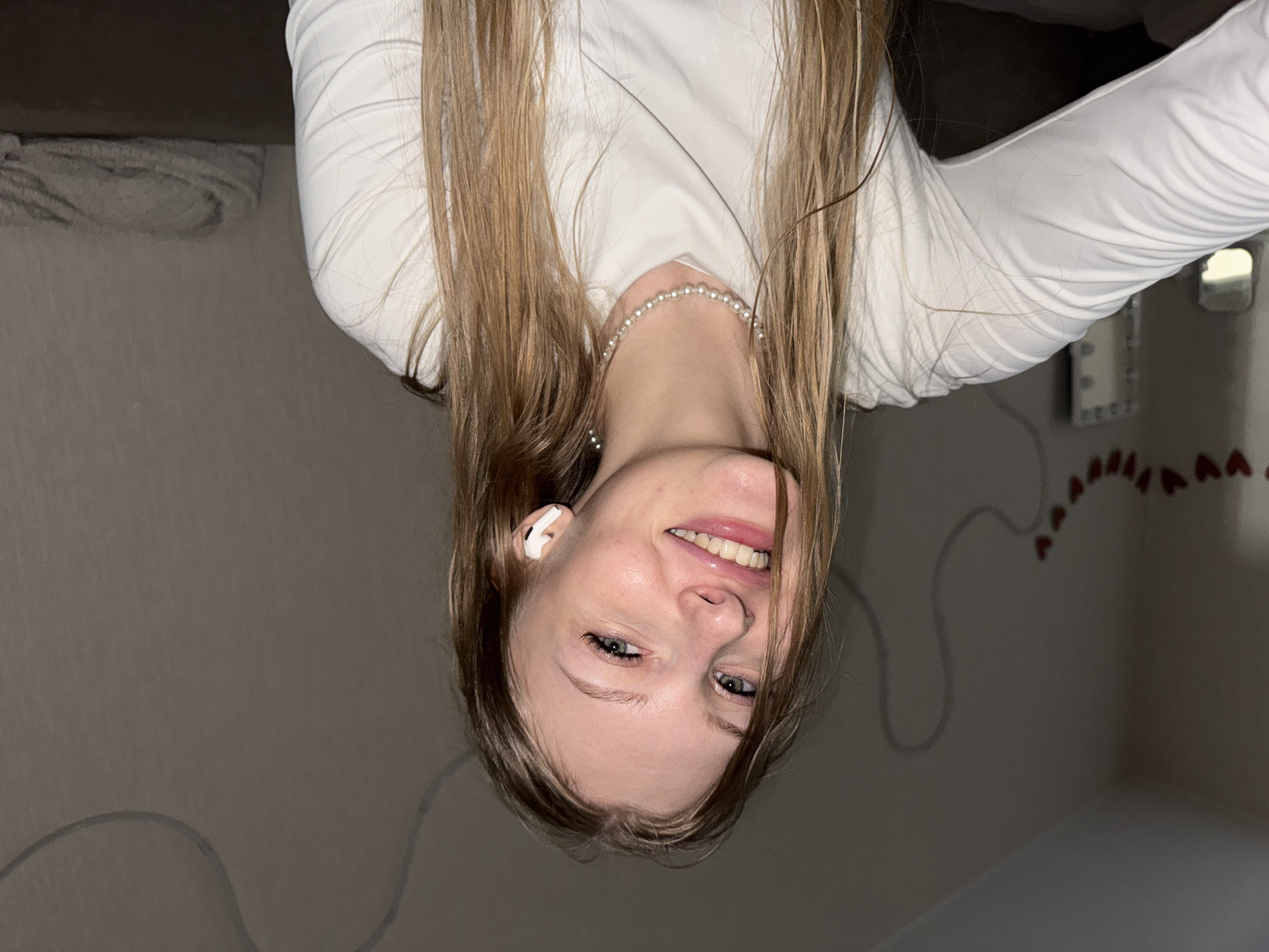 The media file presents an upside-down close-up image of a young woman, likely a selfie. She has long, straight light brown hair and is smiling broadly, revealing light-colored eyes. She is wearing a white long-sleeved top and a pearl necklace. A white wireless earbud is visible in her left ear.
The setting appears to be an indoor room, with a plain, light gray wall forming the background. On the right side of the wall, there is a decorative line of small red heart shapes. Faint, dark lines, possibly wires, are also visible on the wall. The lighting is soft and indoor, suggesting the image was taken in the evening or night. The woman's expression conveys happiness and playfulness. No visible text is present in the image. The location context for this image is Perm, Russia.