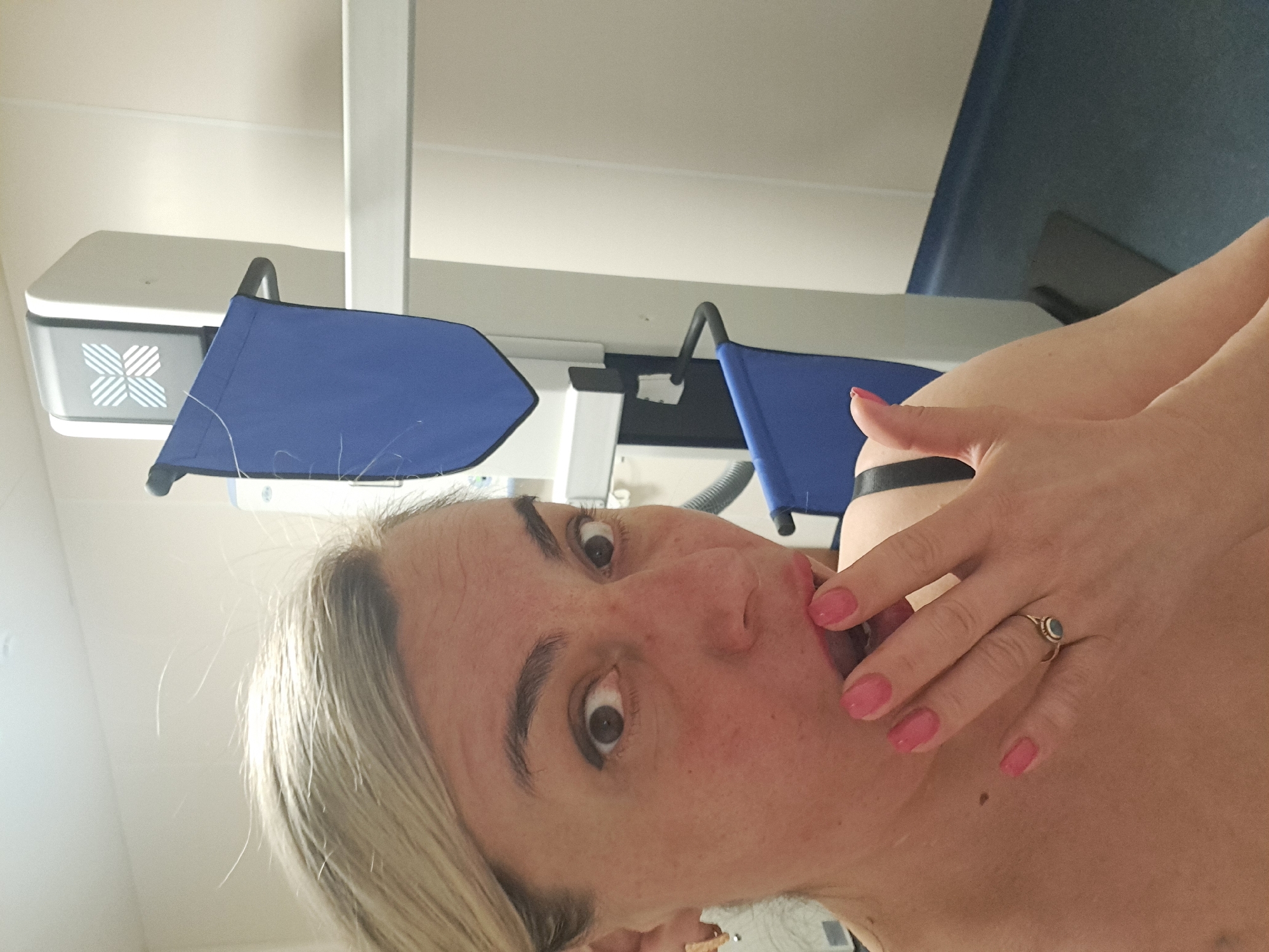 A selfie-style photo shows a woman with blonde hair and pink nail polish, who appears to be indoors in what looks like a medical examination room, with part of an X-ray or mammogram machine visible in the background. The location is not identifiable as Elektrostal, Russia, from the visual information provided. The woman's face is close to the camera, and her hand is near her mouth. The scene is well-lit.