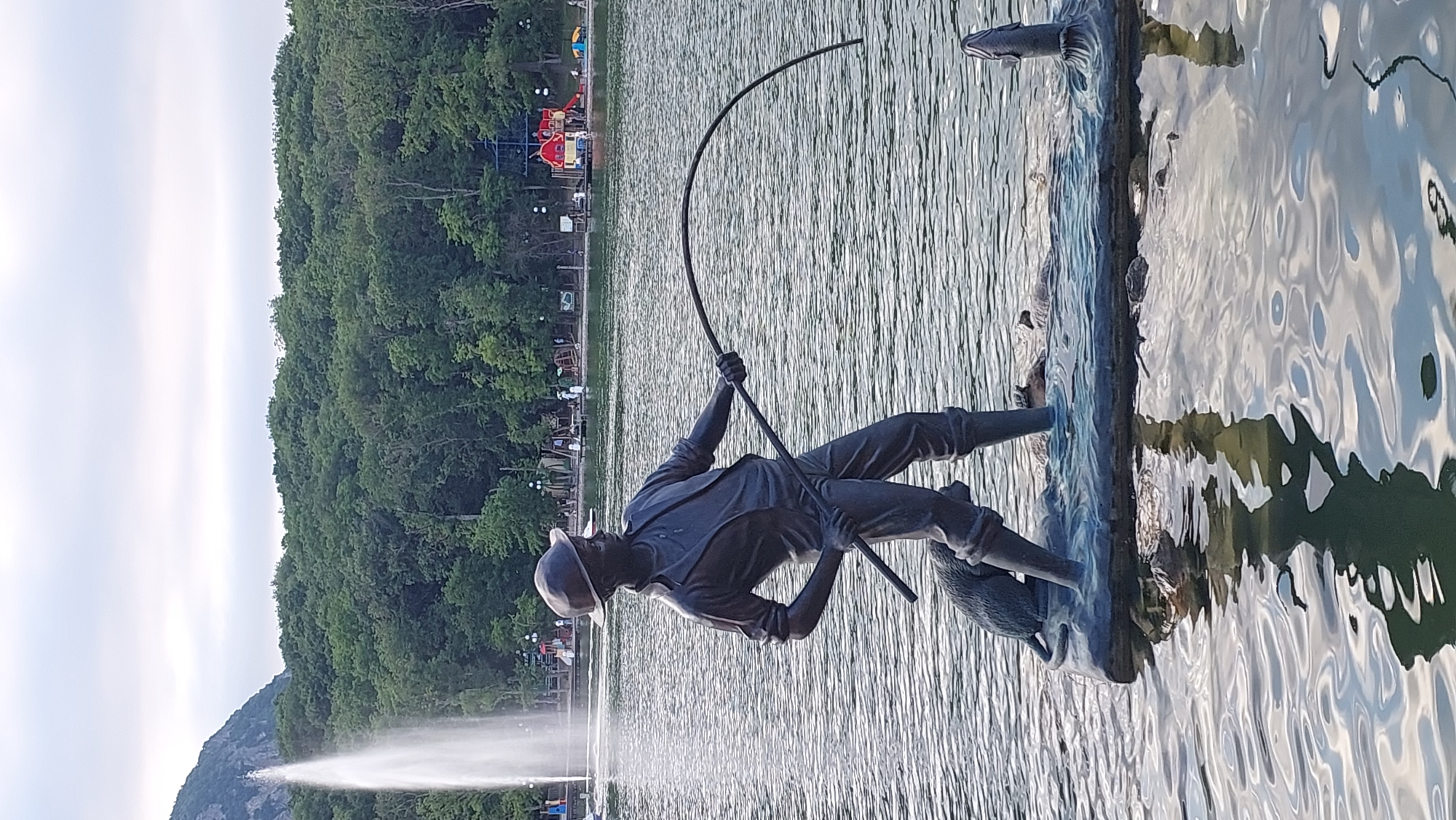 This is a vertical, eye-level, medium shot of a bronze statue of a fisherman in a body of water. The statue depicts a man in a hat and suit, holding a fishing rod that curves upwards, as if he has caught something. He is standing on a stone ledge, with a pelican or similar bird statue at his feet. The water is rippled and reflects the sky and surrounding trees. In the background, a dense green forest rises up a hill, and in the far distance, there appears to be a playground with colorful structures. A faint mist from a fountain is visible in the lower left portion of the image. The lighting suggests it is daytime, with bright sunlight. The scene is tranquil, with the focus on the statue and the reflective water. There is no visible text in the image. The context provided identifies the location as Zheleznovodsk, Russia.