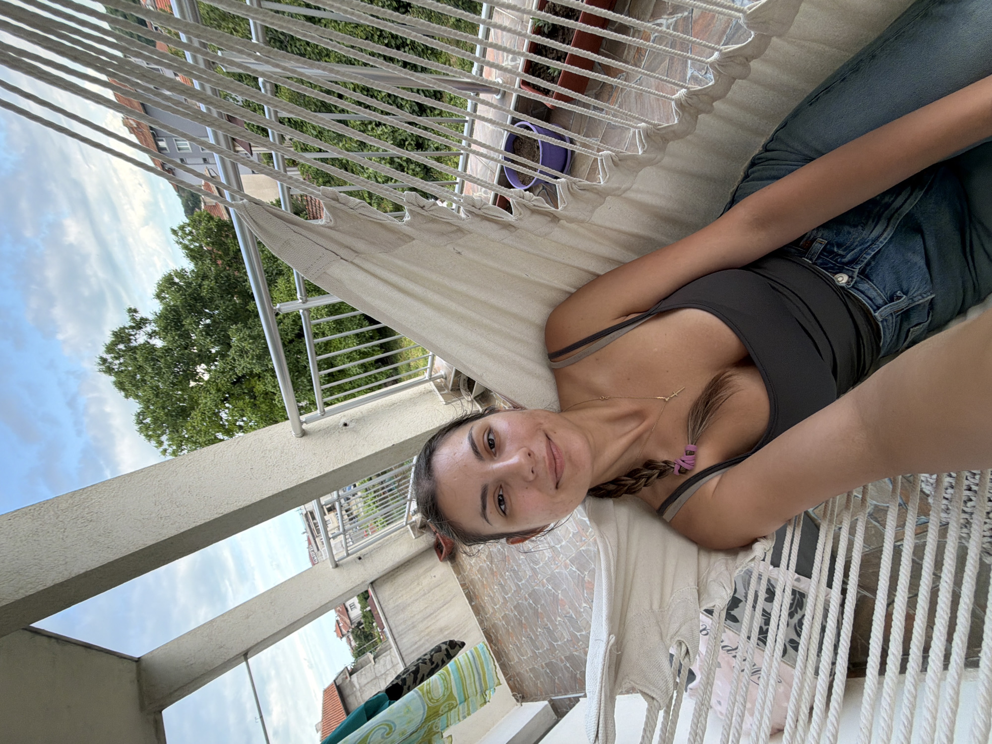 A young woman is lying in a hammock on a balcony in Tsarevo, Bulgaria. The woman has dark hair, braided and tied with a purple scrunchie. She is wearing a dark gray tank top and blue jeans. Her gaze is directed at the camera. In the background, there is a railing, houses, trees, and a cloudy sky. The balcony floor is tiled with a pattern of pink and brown stones. There are two potted plants on the balcony, one in a purple pot and the other in a brown rectangular pot. Clothes are hanging on a rack to the left of the woman. The hammock is made of white woven fabric and ropes.