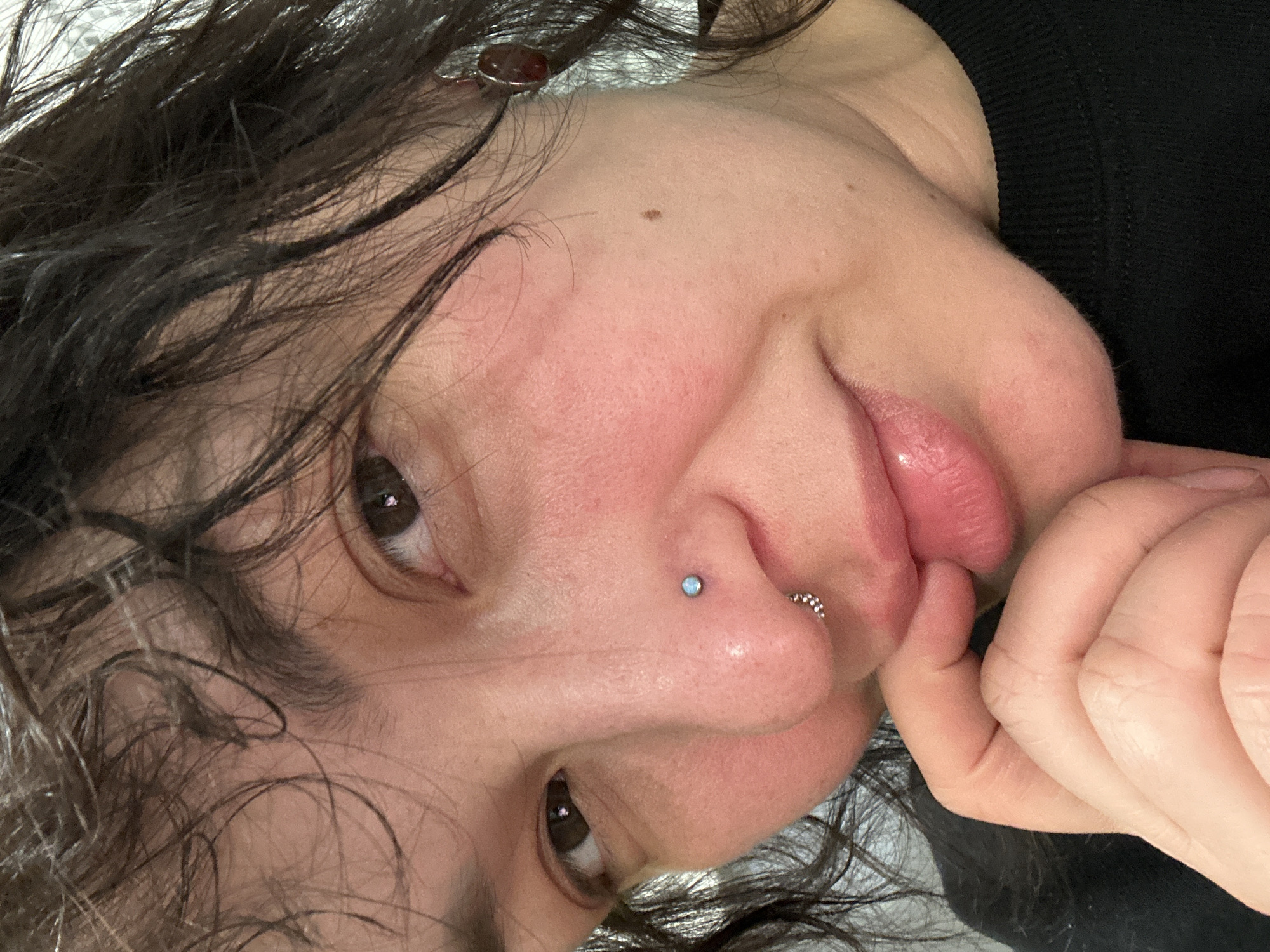 A close-up, tilted portrait shows a person with dark, damp curly hair. The person has a fair complexion with some redness on their cheeks. They have brown eyes and several piercings on their nose: one with a light blue stud and another with a ring. They are touching their lip with their fingers. They have a mole on their forehead. The person is wearing a black garment. There is no information to confirm the location is in the city of Chaikovsky, Russia.
