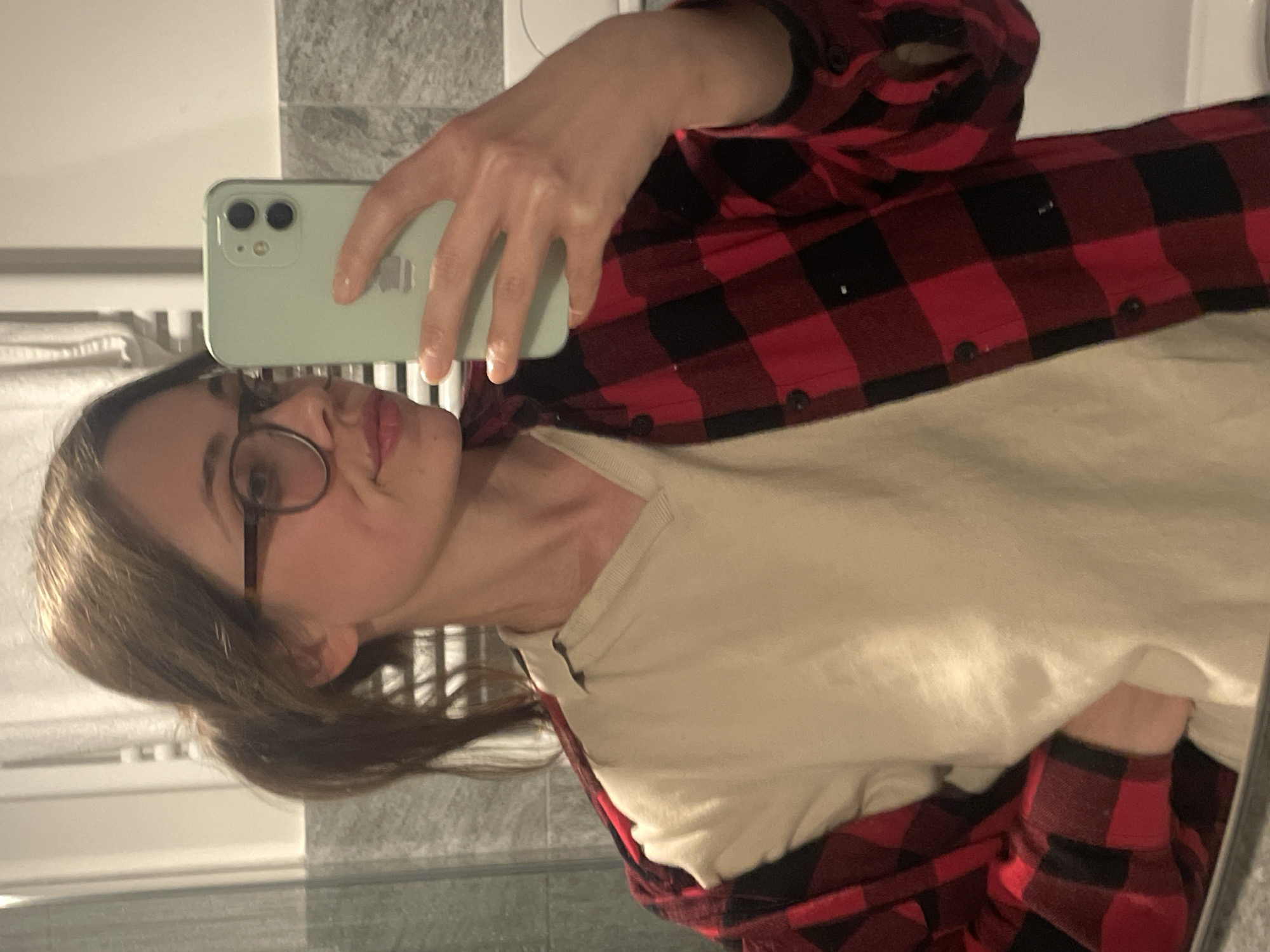 A person with glasses is taking a selfie in a bathroom. They are wearing a white shirt with a red and black plaid flannel over it. The phone is light green. The background is a light gray tiled wall. The location is not discernible as Sappada / Plodn / Sapade, Italy based on this image.