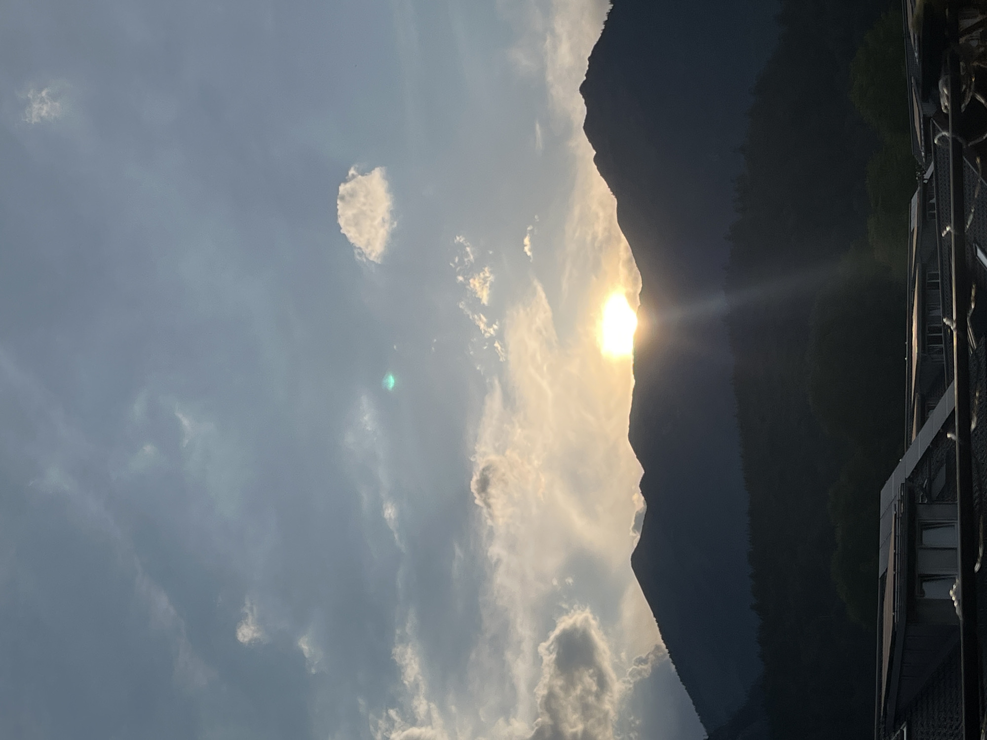 The image depicts a sunset over the mountains in Garmisch-Partenkirchen, Germany. The sun is a bright orb peeking through the clouds, casting a golden glow on the sky and the peaks of the dark, silhouetted mountains. The foreground shows a section of rooftops, possibly of houses or buildings, with some solar panels visible on one of them. The sky is a mix of blues and grays, with scattered clouds that are illuminated by the setting sun. The overall atmosphere is serene and picturesque, capturing the natural beauty of the Bavarian Alps at dusk.