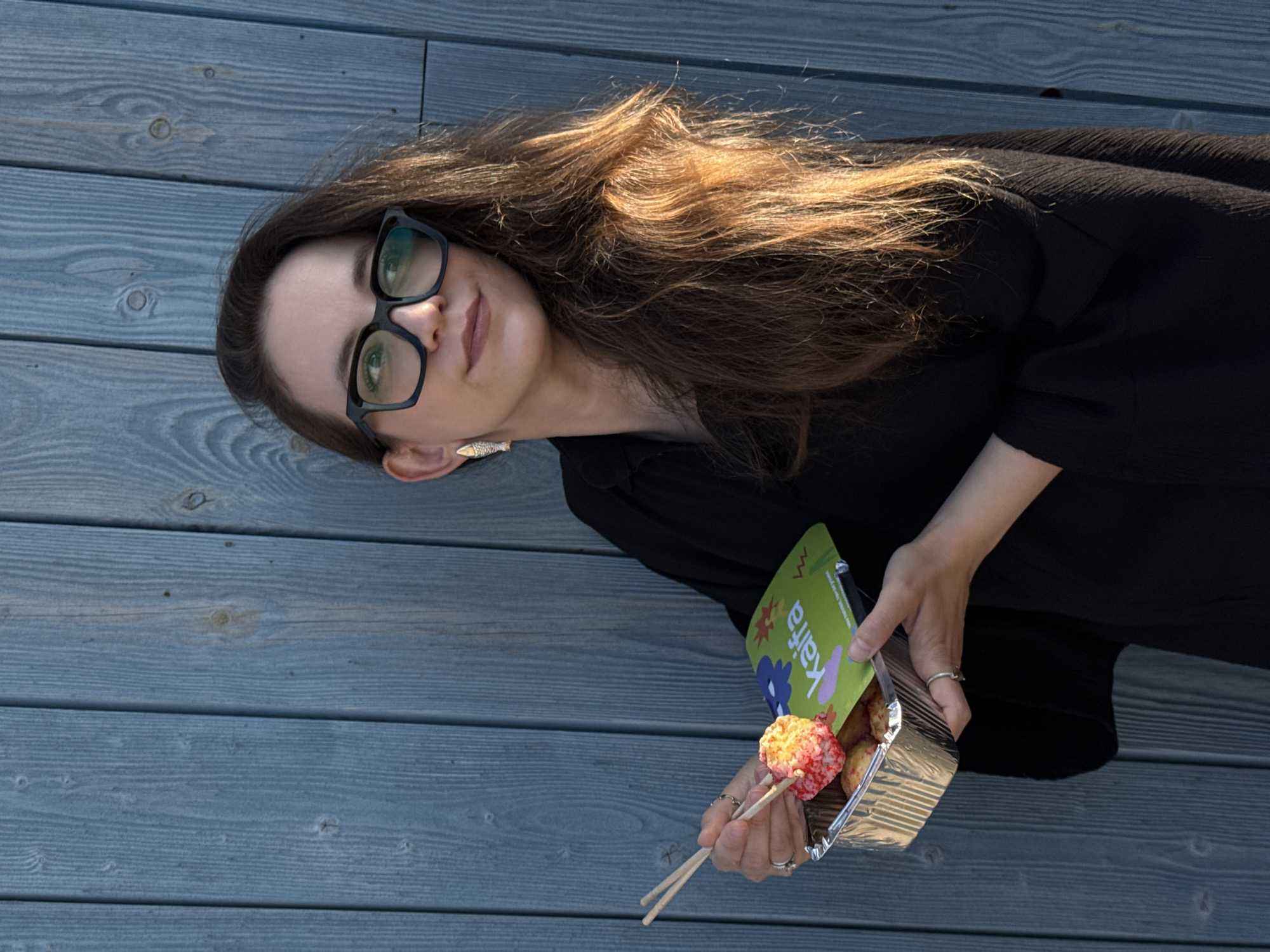An adult Caucasian woman with long, brown hair and black-rimmed glasses is holding a foil container of food. She is wearing a black top and a gold fish-shaped earring. She is using chopsticks to pick up a piece of food from the container. In front of her is a green card with the word "kaifa" written on it in colorful letters. The woman is standing in front of a blue wooden fence. The image was taken in Chaikovsky, Russia.