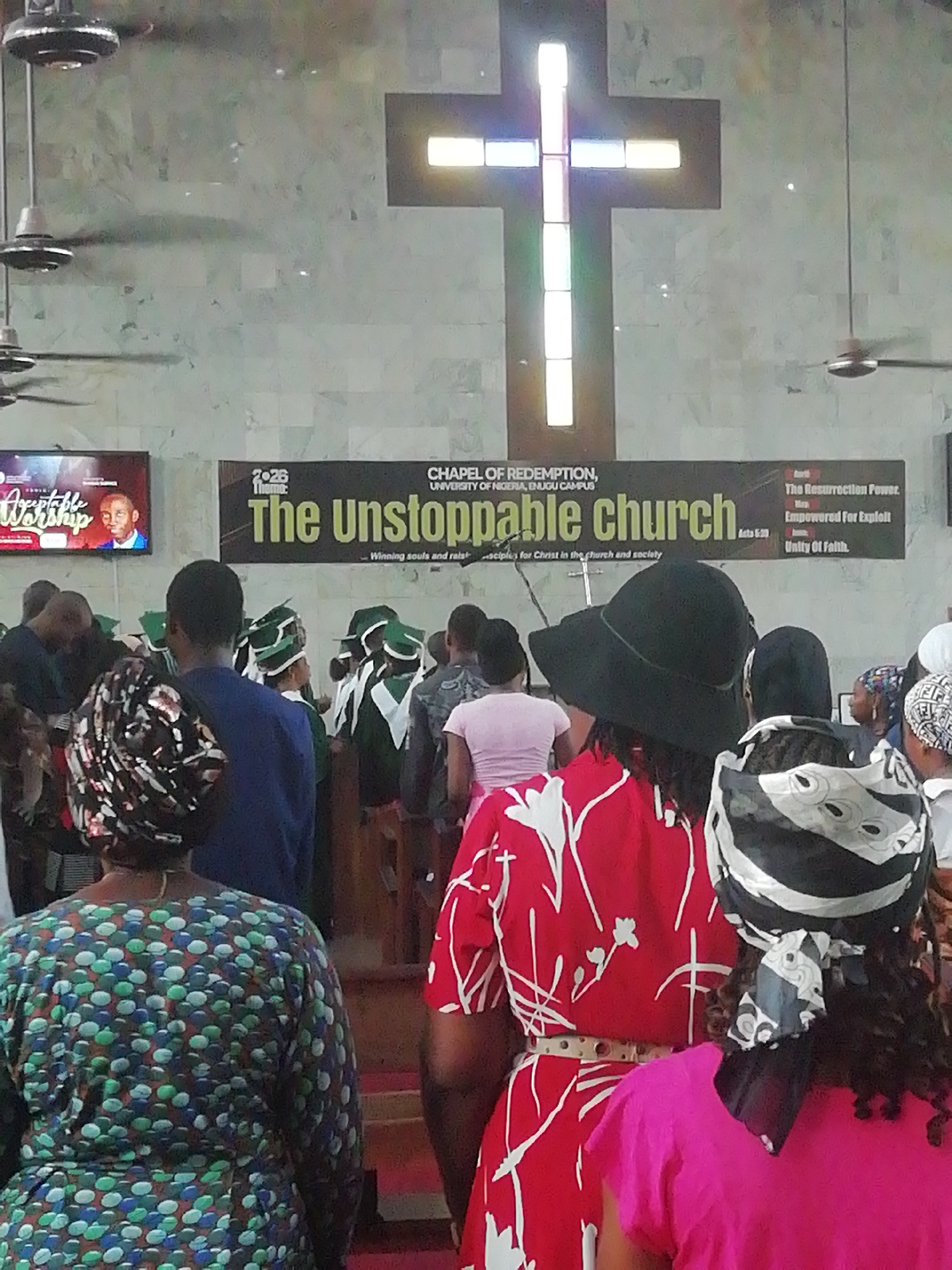 The image shows the interior of the Chapel of Redemption at the University of Nigeria, Enugu Campus, in Enugu, Nigeria. A banner at the front reads "The Unstoppable Church." A large, illuminated cross is prominently displayed on the wall above the banner. Several people are standing and facing forward, likely attending a service. Some individuals are wearing head coverings. A television screen displaying a man and the word "Worship" is visible on the left side of the frame. Ceiling fans are present in the upper portion of the image.