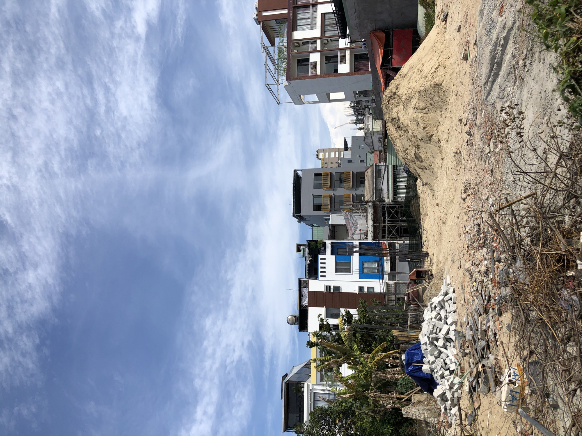 This image depicts a construction site in the city of Đà Nẵng, Vietnam, characterized by a mix of incomplete buildings and debris under a bright, cloudy sky. In the foreground, there are piles of construction debris, including broken bricks, concrete fragments, and branches, with a large mound of sand dominating the center of the frame. A blue tarp is visible covering a pile of white blocks.
The background features a row of multi-story buildings, some of which appear to be newly constructed or undergoing renovation. These buildings exhibit modern architectural styles with balconies, windows, and varying facade colors, including white, brown, and blue. Some structures have rooftop additions, such as terraces or scaffolding. To the right, a building with a white and brown facade and balconies is prominently visible, adjacent to a construction zone with scaffolding and an orange tarp. In the distance, more buildings and construction cranes can be observed, suggesting ongoing development within the urban landscape. The sky is a clear blue, streaked with wispy white clouds, indicative of fair weather. A single person, dressed in dark clothing, is partially visible near the sand pile, appearing to be working.