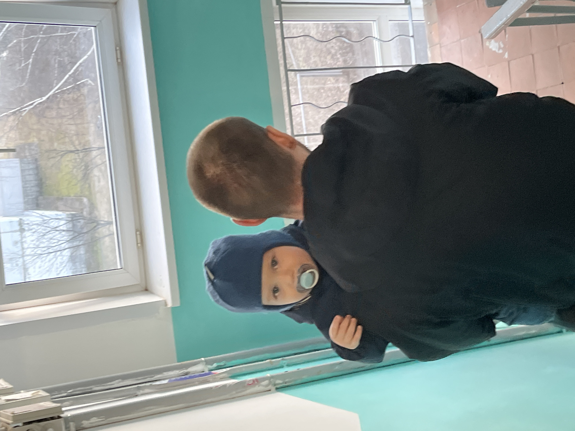 A man in a black hooded sweatshirt is holding a baby in a blue hooded outfit. The baby has a pacifier in its mouth and is looking at the camera. They appear to be inside a building with a window on the left showing a view of trees and a building outside. There is another window to the right with a metal security grate. The wall in the background is a light turquoise color. The photo was presumably taken indoors at a residence or facility in the city of Chaikovsky, Russia.