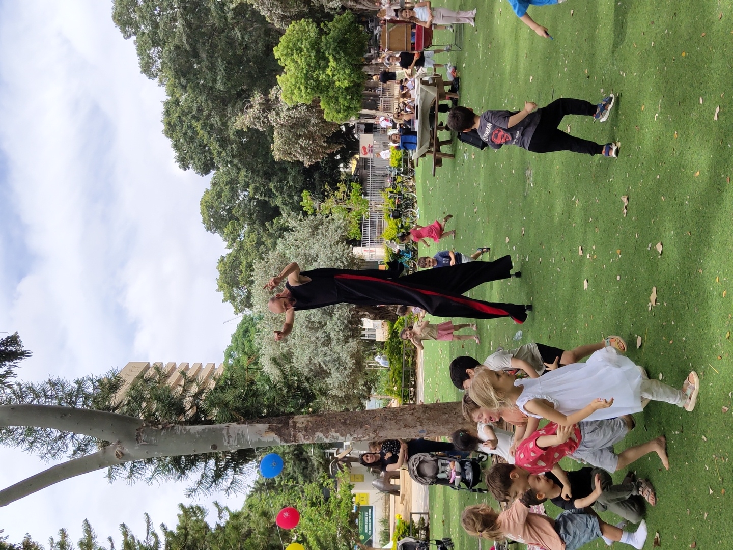 The image shows a group of children gathered on a green lawn, likely artificial turf, in an outdoor setting. One man dressed in a black outfit with red stripes appears to be performing or interacting with the children, who are clustered near the bottom of the frame, some sitting and others standing. A few other people are visible further back, along with what appears to be a park setting with trees and other greenery. In the background, there are benches and some bicycles. Balloons are tied to a tree on the lower left. A building with a unique tiered architecture can be seen behind the tree. Given the urban environment and the general description, the location is likely in a park in Tel Aviv, Israel.