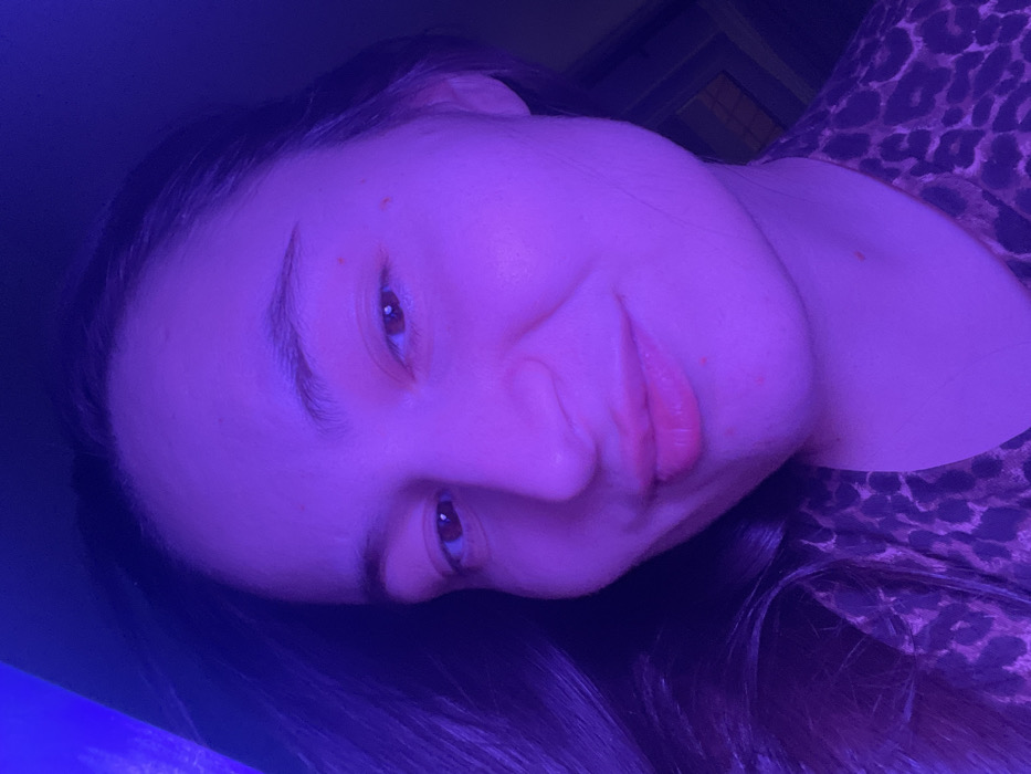 A woman is captured in a close-up, slightly angled shot, bathed in a blue light that tints her skin and surroundings. She has fair skin, dark hair, and light-colored eyes. Her expression is soft, with a slight smile. She's wearing a patterned top, possibly animal print. A faint glow appears on the lower left of the image, likely the source of the blue light.