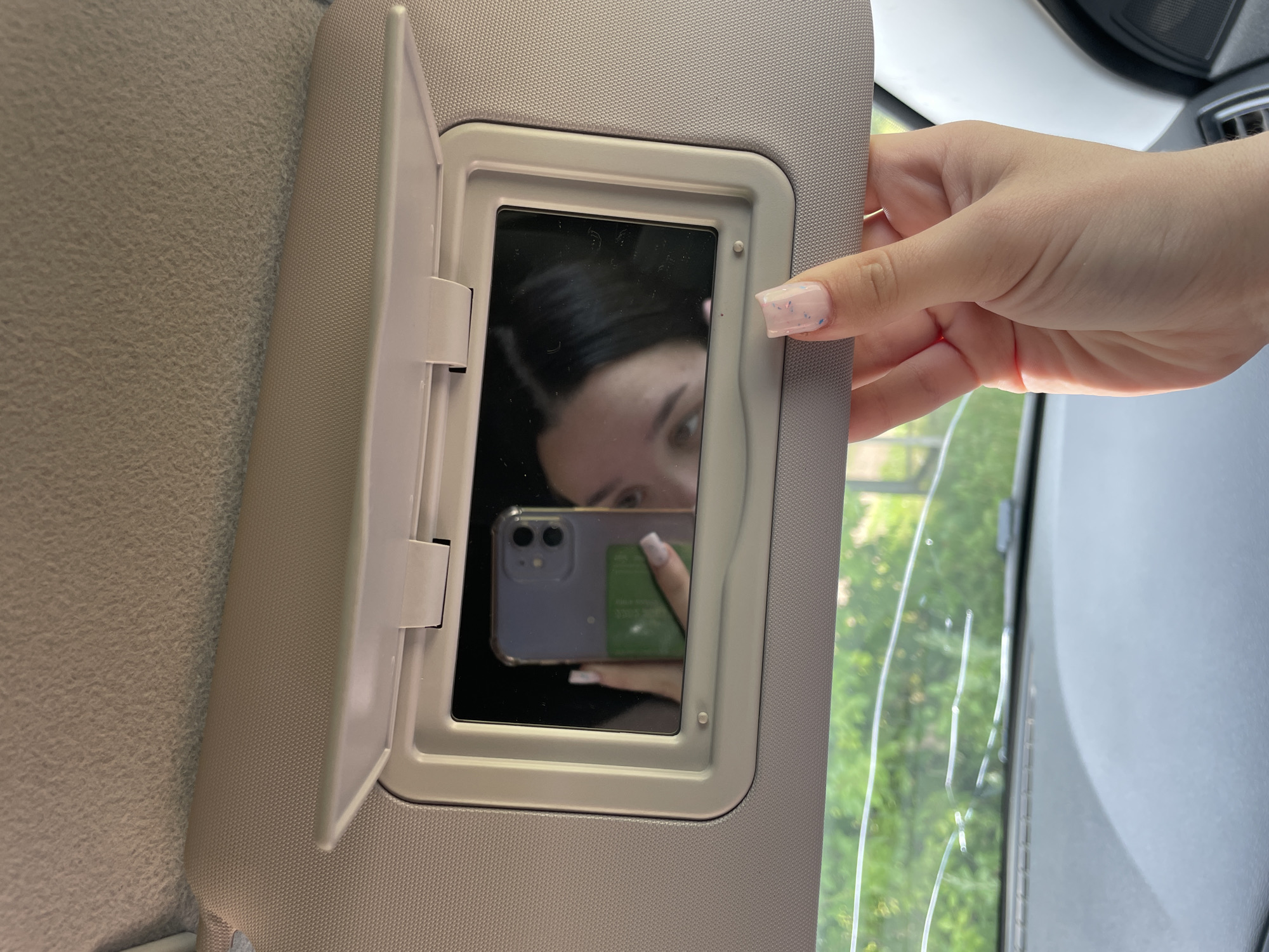 In this close-up shot, a person's hand, adorned with light pink nail polish speckled with blue, is visible in the upper right quadrant of the image. The hand is interacting with a car's visor, which is open to reveal a mirror. The mirror reflects a person's face and a smartphone. The person in the reflection has dark hair and is looking towards the phone. The phone itself is a light purple color with a dual-lens camera. A green item, possibly a card with white text, is partially visible behind the phone. The background visible through the car window suggests an outdoor setting with lush green foliage, implying it is daytime and likely pleasant weather. The scene appears to be taking place inside a vehicle, given the presence of the car visor. No specific text is clearly legible beyond what might be on the green card. The overall mood is casual, possibly a moment of checking one's appearance or taking a quick selfie.