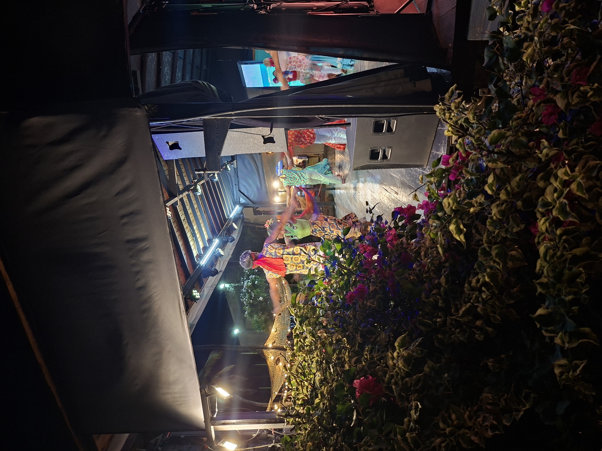 The image depicts an outdoor evening performance stage in Porlamar, Venezuela, likely a cultural event. In the foreground, lush green foliage with pink and purple flowers frames the scene, obscuring the lower portion of the stage. The stage itself is made of wood and is illuminated by spotlights and decorative string lights, creating a warm and festive atmosphere. Several performers, dressed in brightly colored, traditional attire, are dancing in a circle. One individual, wearing a yellow and white patterned shirt and a red headscarf, is prominent in the center of the frame, with arms outstretched. The background reveals more tropical vegetation and palm trees against a dark night sky. Large black speakers are positioned on the sides of the stage. The overall impression is one of a lively cultural celebration taking place in a tropical setting.