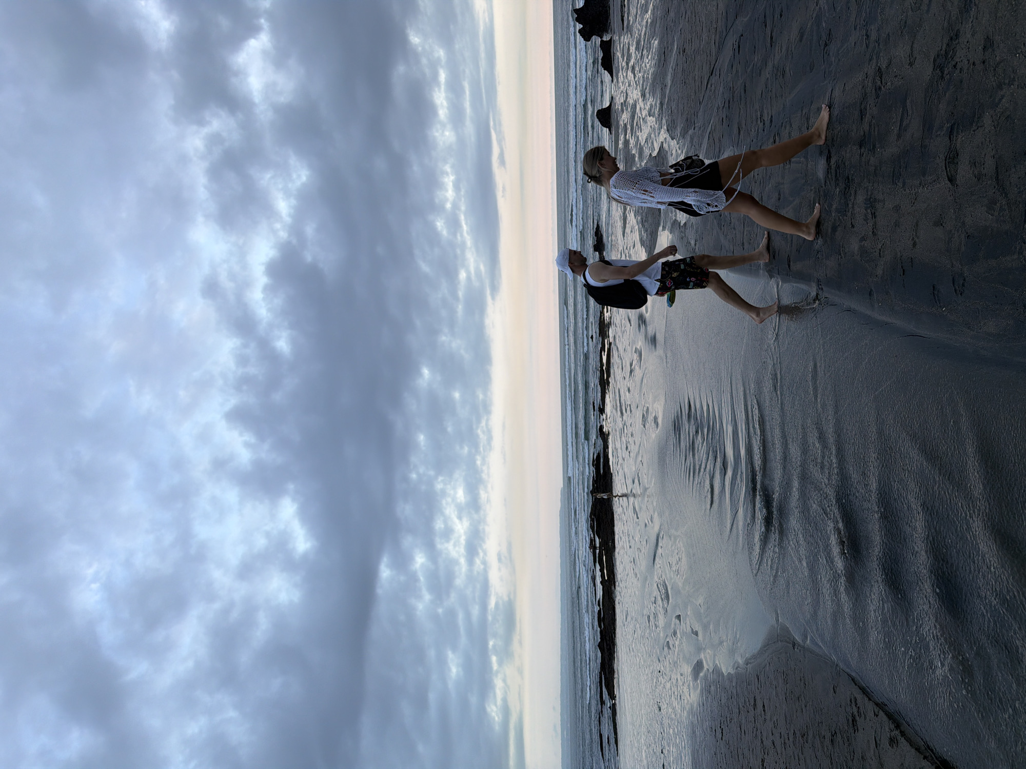 Two people are walking along a black sand beach at sunset in North Kuta, Indonesia. The sky is overcast with grey clouds. The sea is calm with small waves breaking on the shore. The beach is wet and reflects the sky. The person in the foreground is wearing a white shirt, dark patterned shorts, and a white cap, with a black backpack. The person in the background is wearing a white crochet top and dark shorts. They are walking away from the viewer. There are dark rocks or debris along the shoreline.