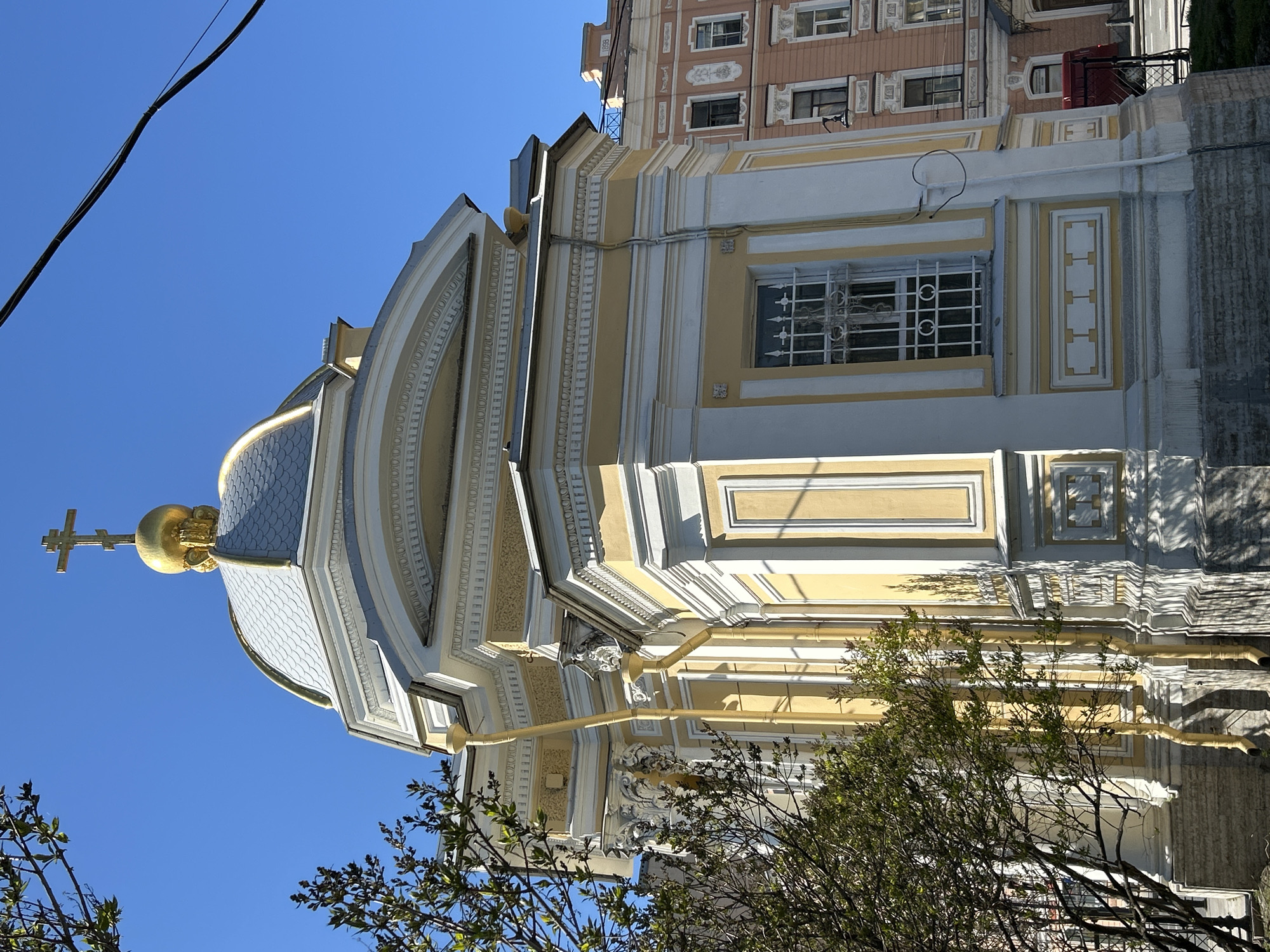 The image shows a section of a yellow and white building, likely a church, with a blue and silver dome topped with a golden cross. The building has ornate details and a window with a decorative iron grate. A building with multiple windows is visible behind the church. A tree with green leaves is in the foreground. A power line is visible in the upper left corner. The clear blue sky indicates a sunny day. The building is located in the city of Saint Petersburg, Russia.