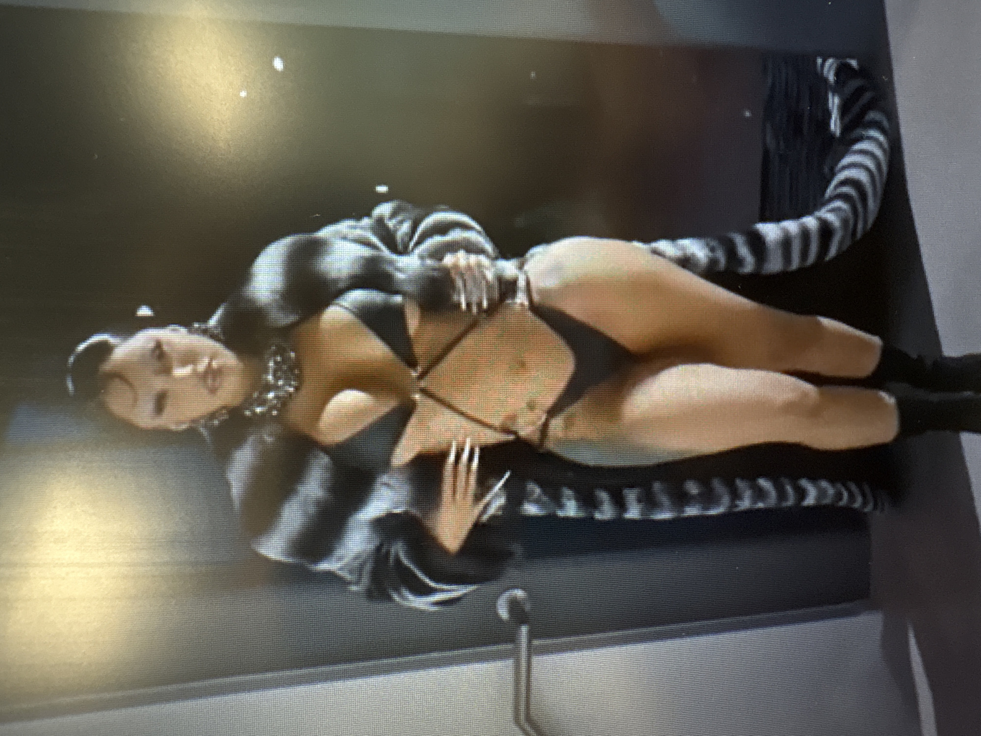 A woman stands facing the camera, wearing a black bikini, a large, fluffy, black and white striped coat, and black socks. She has a large, silver necklace around her neck and her hair is styled in a high bun with curls framing her face. She is standing in what appears to be an indoor setting with a gray wall and a door to her left. The lighting is bright and casts shadows on the wall behind her. No specific location in Sofia, Bulgaria can be determined from the image.