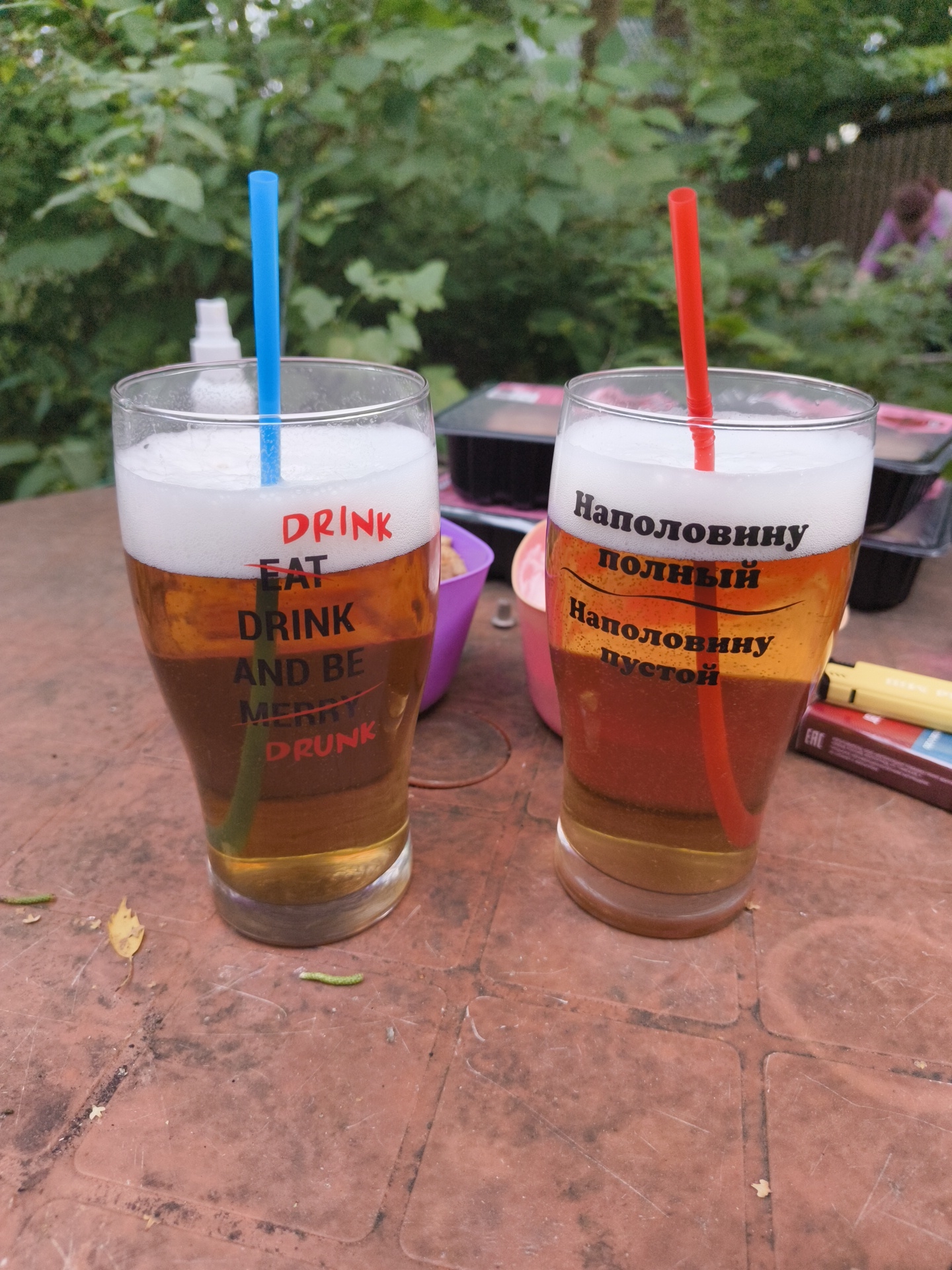 Two glasses of beer with straws are on a red plastic table outdoors. The glass on the left has red text that says "DRINK EAT DRINK AND BE MERRY DRUNK". The glass on the right has Russian text. In the background, there are green trees and a wooden fence. There are also some food containers and other objects on the table. The overall scene appears to be a casual outdoor gathering.
