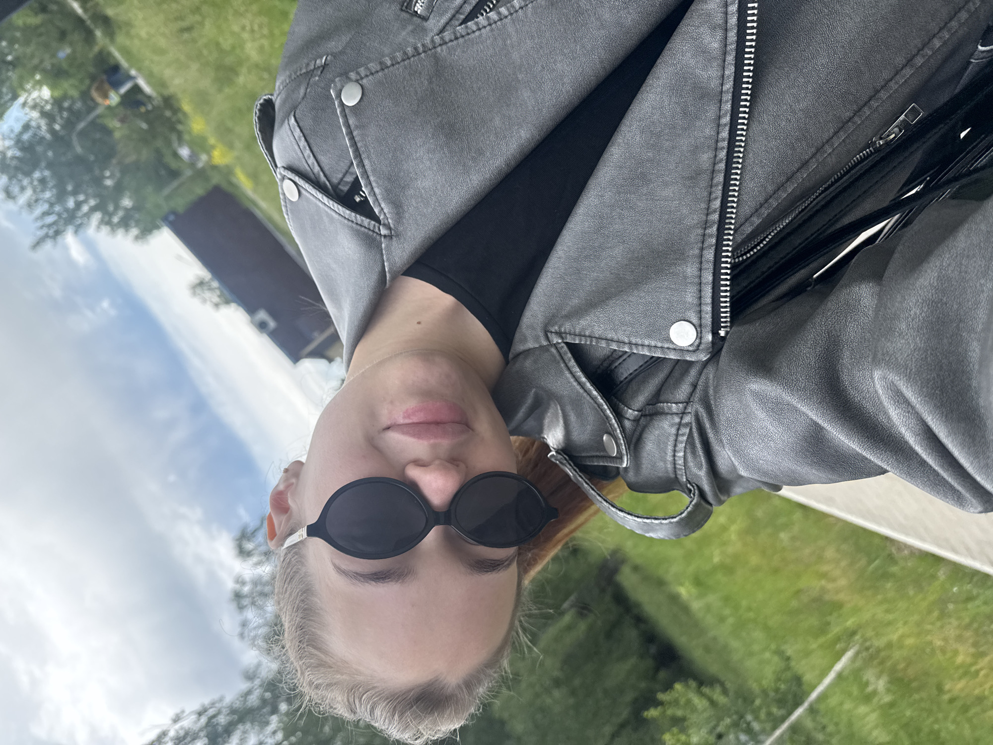 A person wearing sunglasses and a grey leather jacket poses for a selfie outdoors. The background shows a green grassy area, trees, a building with a brown roof, and a cloudy sky. I am unable to determine the specific location within Chaikovsky, Russia.