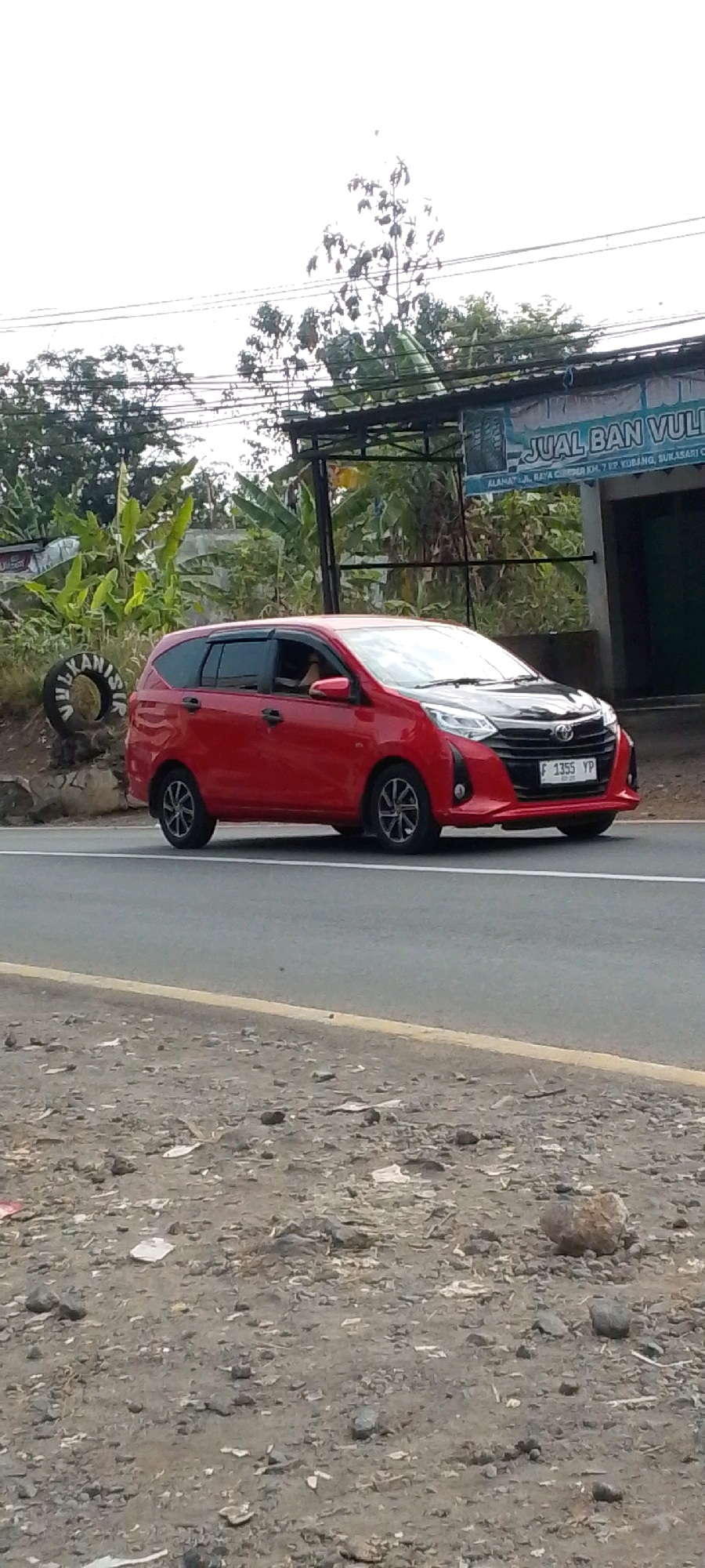 A red car with a black hood is parked on the side of a road. The car has the license plate F 1355 YP. In the background, there is a building with a sign that says "JUAL BAN VULCANIZING" which translates to "Vulcanized Tire Sales." This business is located on Jalan Raya Cibunar, Km. 7, Cipatat, Sukasari, Cilaku, Indonesia. The scene appears to be in a rural or semi-urban area, with trees and foliage visible. The ground is covered with gravel and debris. There are no people clearly visible in the image.