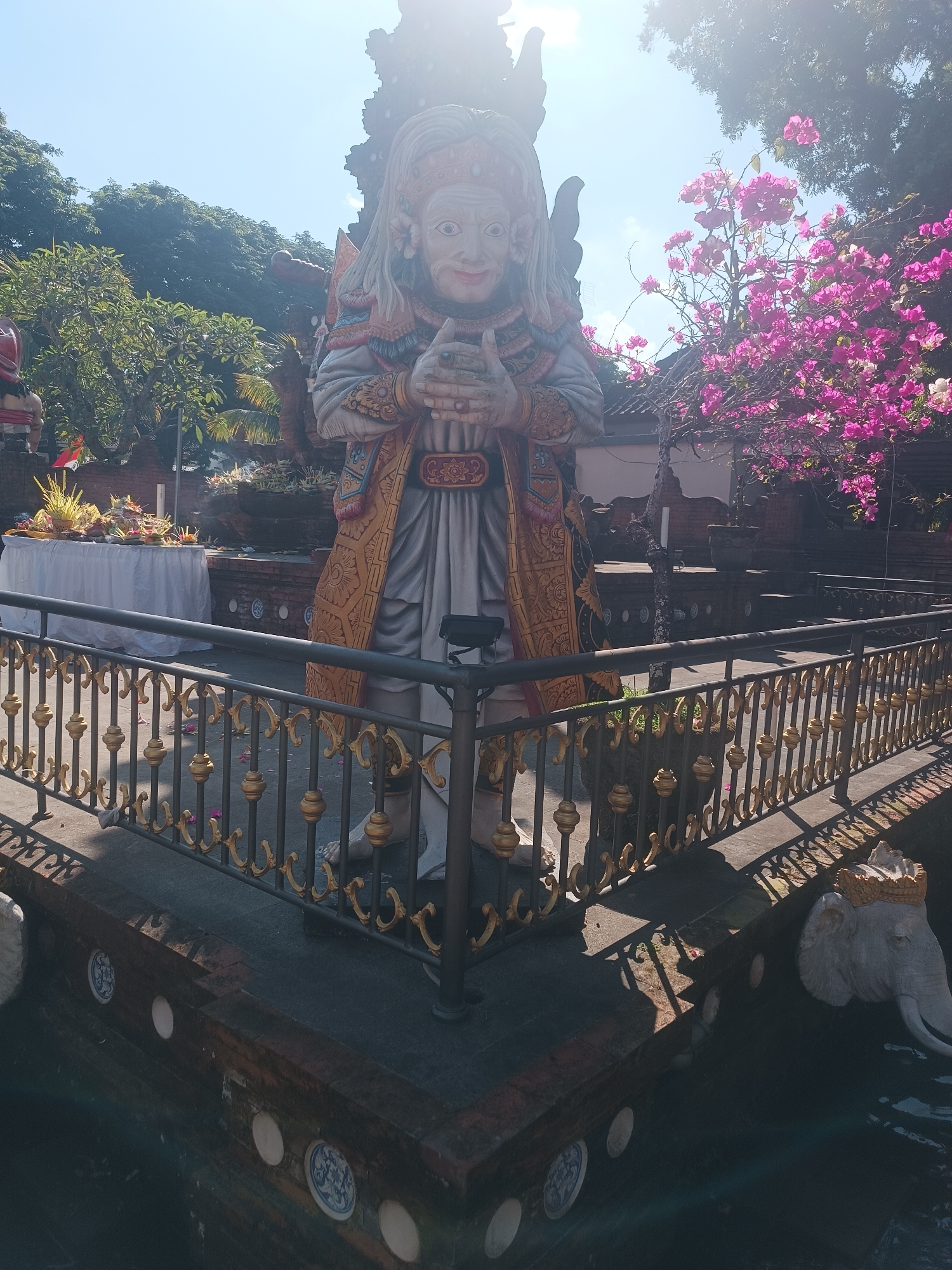 A full, slightly low-angle outdoor shot depicts a large, traditional Balinese statue at the Pura Taman Ayun, a royal temple in the village of Mengwi, Badung Regency, Bali, Indonesia, near the city of Tabanan. The statue is a character from Balinese mythology, adorned with a gold and red headdress, elaborate gold-patterned robes, and detailed facial features, with its hands clasped in front. The statue stands on a dark, ornate stone platform. In the background, lush green trees, a bright blue sky, and a vibrant pink bougainvillea bush are visible. In the middle ground, a white-covered table with offerings of food and flowers, and a brick wall with carved decorative elements can be seen. A black metal fence with gold accents surrounds the foreground area, and the lower part of the frame shows a dark water feature with a stone elephant head sculpture. The scene is illuminated by bright sunlight, casting shadows on the ground and the surrounding structures.