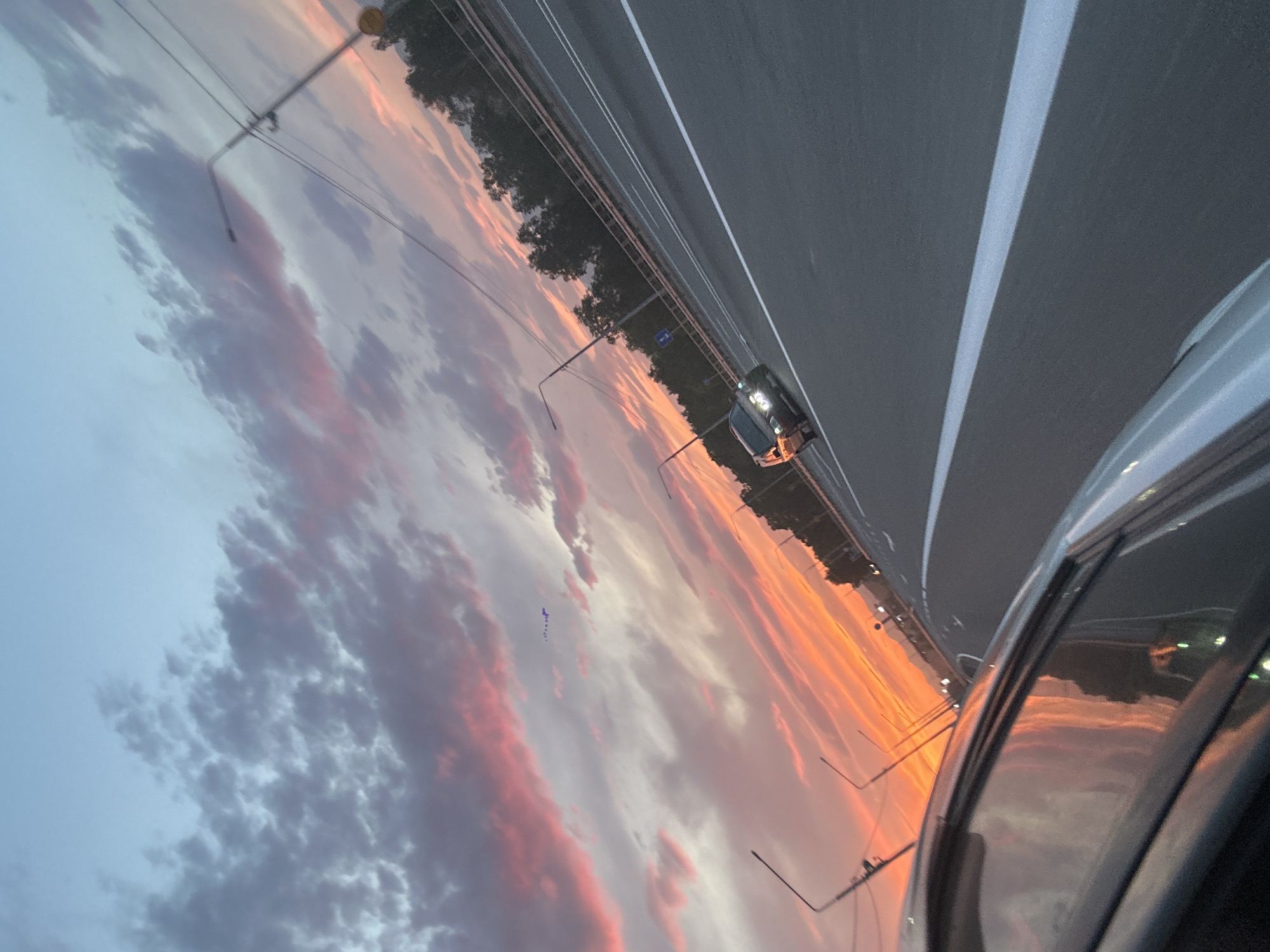 A photograph captures a road and the sky at sunset, as viewed from inside a vehicle. The sky is filled with clouds illuminated by the setting sun, displaying vibrant hues of orange, pink, and purple against a backdrop of pale blue. The road is asphalt, with white lane markings visible. On the right side of the road, there is a guardrail and a line of trees. Streetlights line the left side of the road. A dark-colored car is driving on the road in the middle distance. The lower left portion of the image shows the interior of a vehicle, with the side of the car and its reflection of the sunset visible. The scene appears to be on a highway or a major road, possibly on the outskirts of Yelizovo, Russia, given the overall landscape and the presence of infrastructure typical of such a location.