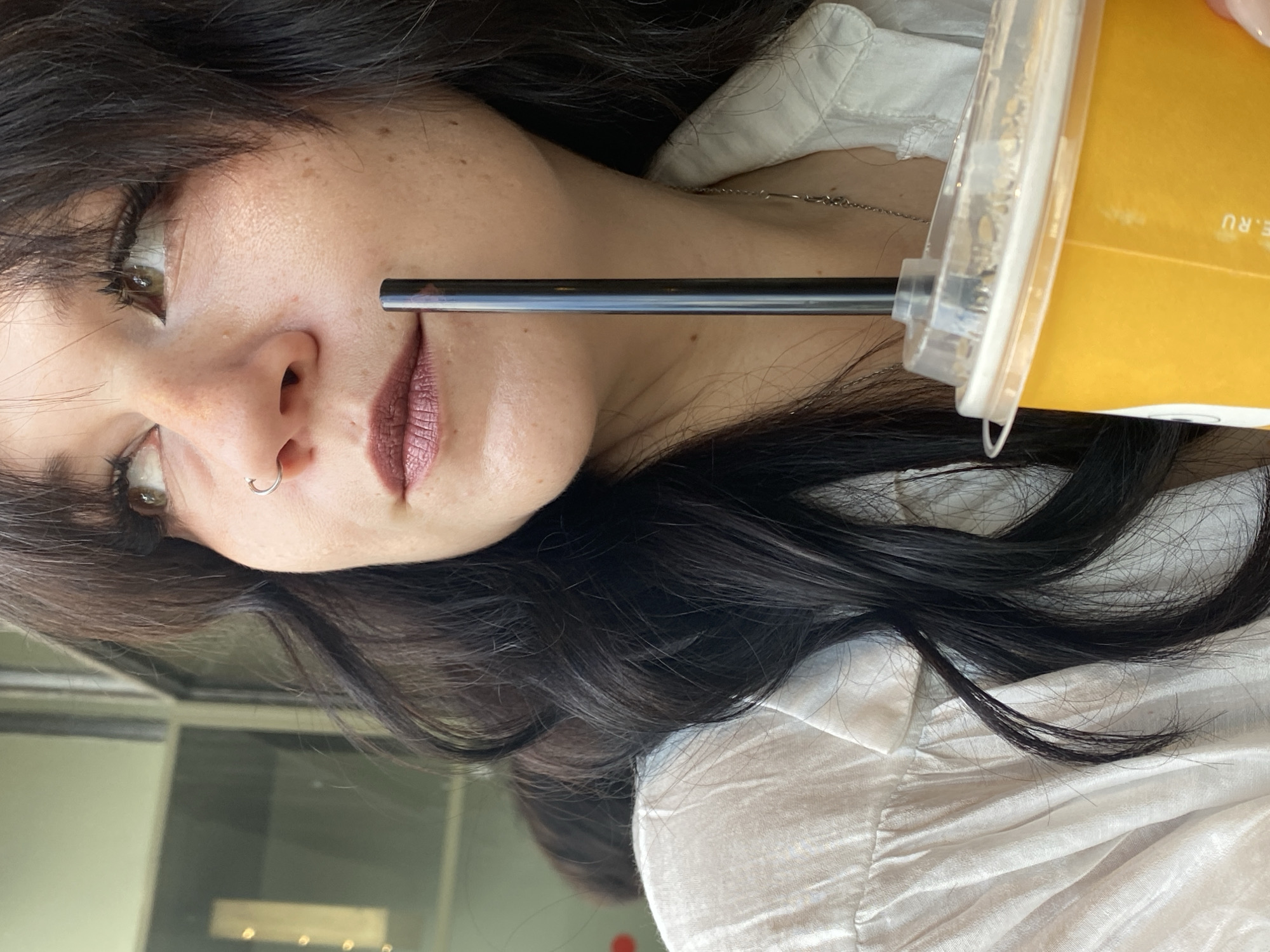 A woman with dark hair and a nose ring, wearing a white shirt, holds a cup of orange liquid with a black straw. She is looking up and to the right. The background is out of focus, showing what appears to be a window with reflections and a glimpse of a building facade. The overall scene suggests an indoor or shaded outdoor setting, potentially a cafe or restaurant. It is not possible to determine the specific location within Saint Petersburg from the provided image.