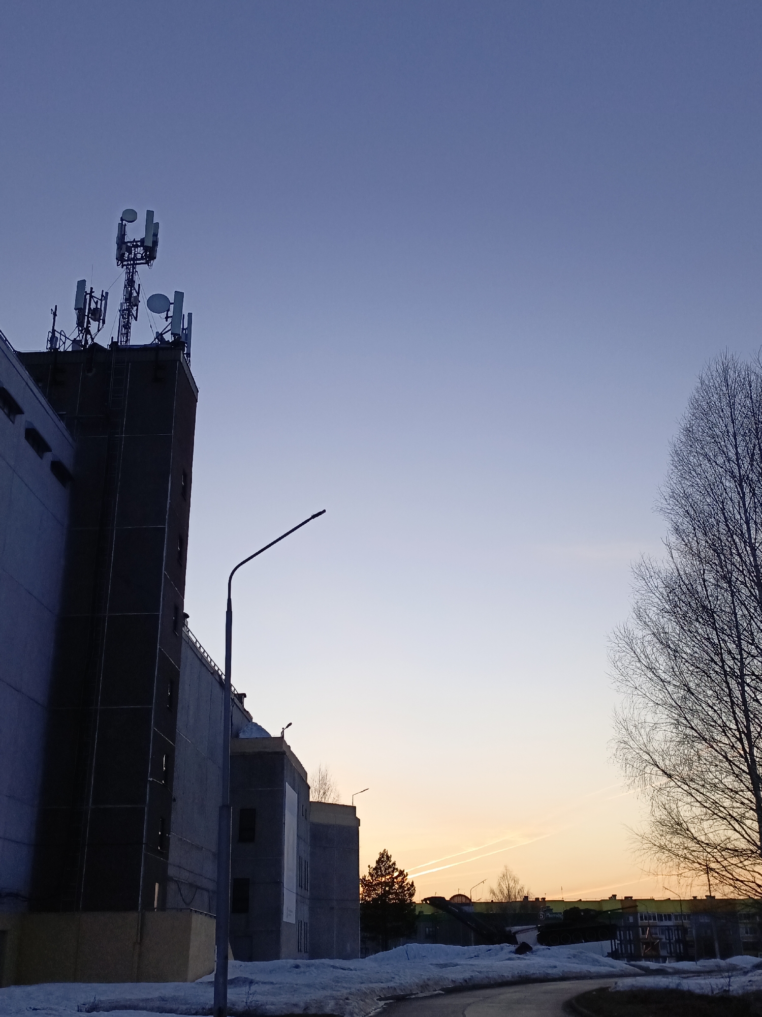 The image shows a view in Марковский, Russia, with a building dominating the left side of the frame. The building has several cellular communication antennas on its roof. A street lamp stands in front of the building. There is snow on the ground. In the background, there are more buildings, trees, and a road. The sky is transitioning from blue to orange, indicating sunrise or sunset.