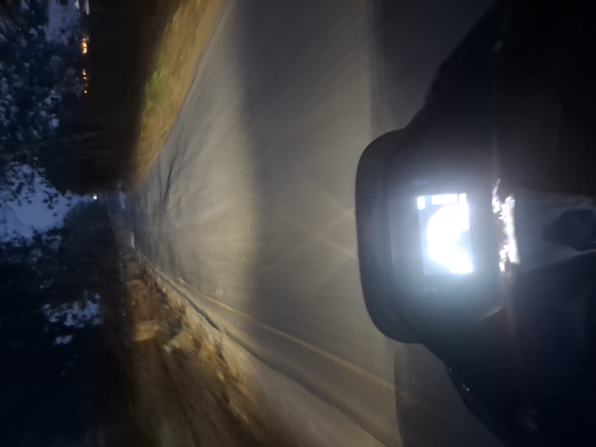 The image captures a first-person perspective from a vehicle, likely a motorcycle, traveling on a paved road at dusk or night. The road has yellow lane markings and is illuminated by the vehicle's headlights. The surrounding environment appears to be rural or suburban, with trees and foliage visible on either side of the road. In the foreground, a digital display screen from the vehicle is prominent, emitting a bright light. The screen's content is not clearly discernible due to overexposure and blur. The sky is dark, with some faint light suggesting the transition from day to night. There are no visible people or other vehicles in the immediate vicinity. The location is identified as Gampang Sejati, Indonesia.