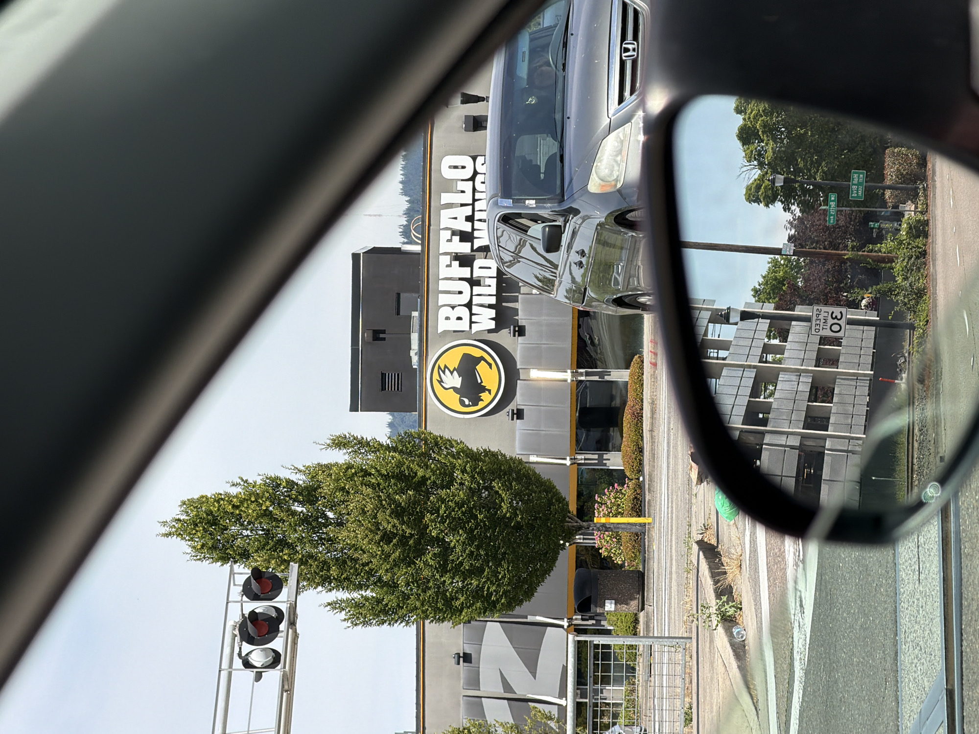 The image shows a view from inside a vehicle looking out towards a Buffalo Wild Wings restaurant. In the foreground, a car is visible, and the side mirror reflects a street scene with buildings and trees. Outside the car, a tall evergreen tree stands in front of the Buffalo Wild Wings building. The restaurant has a prominent sign with its logo and name. To the left of the tree, a metal fence encloses a sidewalk area with a trash can. Traffic lights are visible in the upper left portion of the image. The background shows a clear sky and some distant trees. The scene appears to be in a commercial area, likely during daylight hours.