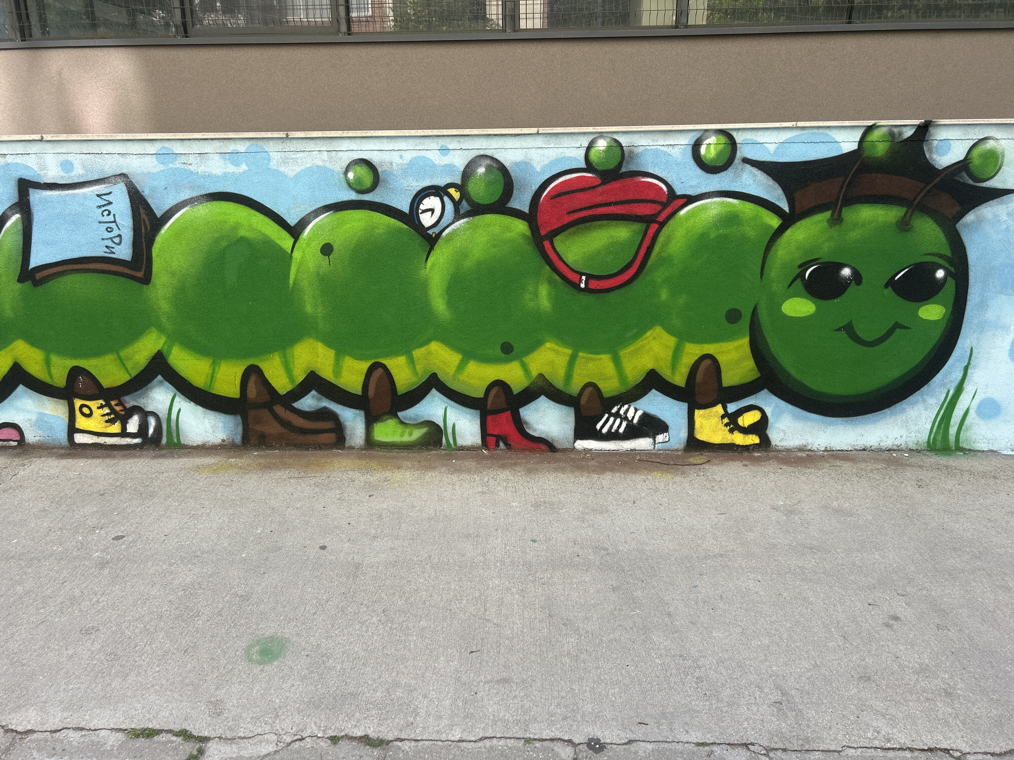 This image captures a vibrant graffiti mural on a light blue concrete wall in Sofia, Bulgaria. The central subject is a large, multi-segmented, cartoonish green caterpillar. The creature is composed of seven distinct, rounded green segments, each outlined in black and featuring yellowish-green shading along its base.
The leftmost segment of the caterpillar displays a light blue rectangular sign with the Cyrillic word "Истори" (Istori) written in black capital letters. The second segment from the left features a small blue and white alarm clock icon near its top. The fourth segment from the left has a bright red, sling-like object or ribbon draped over it. Several smaller green spherical bubbles float above various segments of the caterpillar's body.
The caterpillar's head, positioned on the far right, is round, green, and exhibits a wide, content smile with two large black oval eyes and two small green blush marks on its cheeks. It wears a dark brown or black graduation cap (mortarboard) and has two antennae, each topped with a small green sphere.
Beneath the caterpillar's body, a collection of individual shoes and boots are painted, representing its multiple legs. From left to right, these include a partially visible pink shoe, a yellow high-top sneaker with white laces, a brown boot, a green shoe, a red high-heeled shoe, a black and white athletic sneaker, and a yellow slipper. On the far right, green blades of grass are depicted.
The graffiti is situated on the lower portion of a building. Above the painted wall, a plain brown-grey building facade is visible, featuring several dark-framed windows, some partially obscured by a metal security grid or fence. In the foreground, a grey concrete sidewalk with visible cracks and minimal plant growth extends across the bottom of the frame.