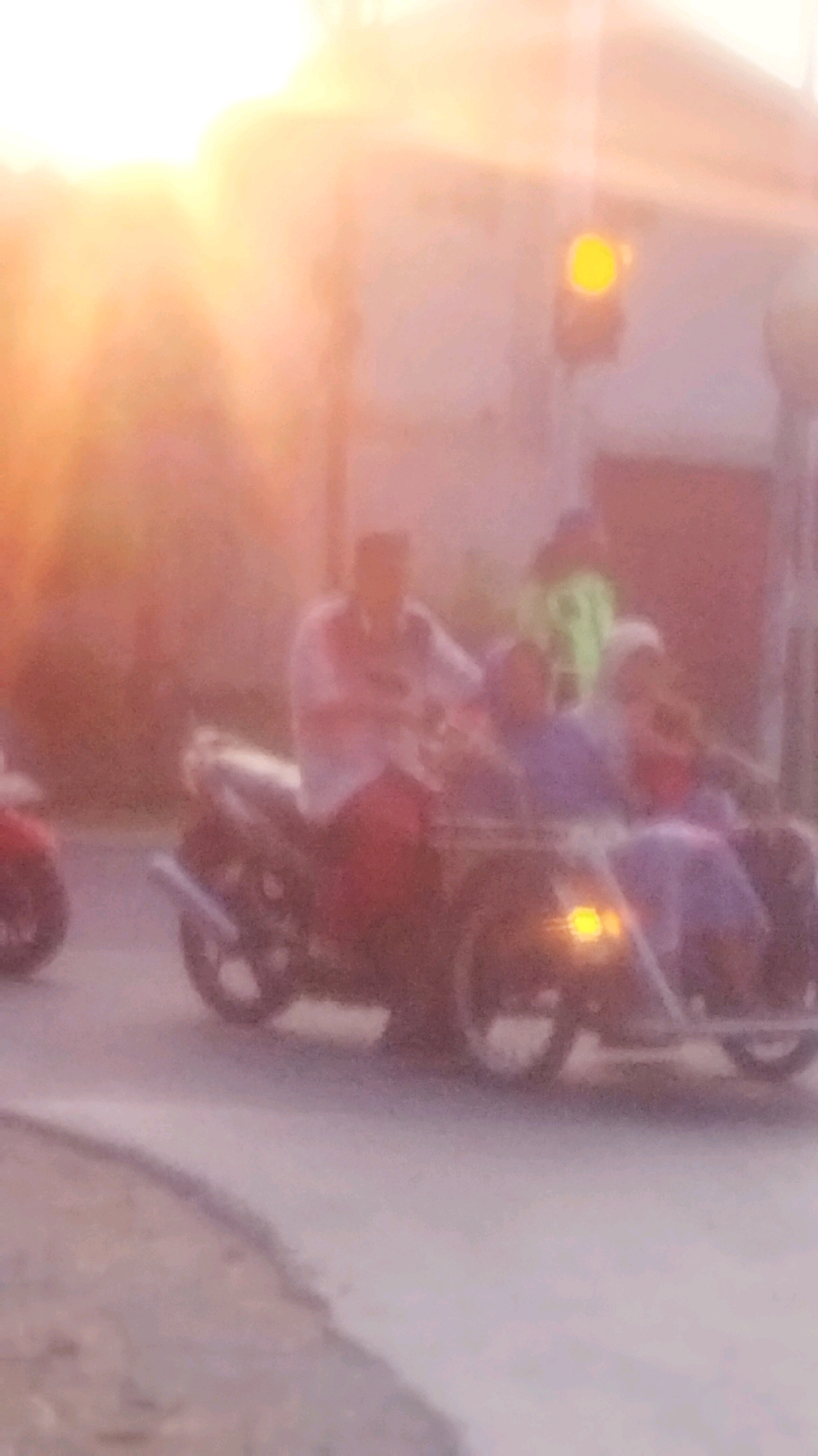 The image captures a scene in Palang, Indonesia, during what appears to be sunset or sunrise, indicated by the warm, hazy light and lens flare. A motorcycle with a sidecar is the central focus, carrying at least three individuals. A person in the front, likely the driver, is wearing a white shirt with red accents and dark pants. Two passengers are seated in the sidecar; one in front wearing a purple top, and another behind wearing a blue top and a headscarf. The motorcycle is on a road, with a traffic light visible in the background, showing a yellow light. Another motorcycle, partially visible on the left, is red. The environment is urban, with buildings suggested in the soft-focus background. The overall impression is one of daily life on the road in Palang, characterized by the warm, diffused light of dawn or dusk.