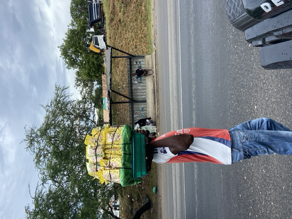 A man carries a large green basket piled high with yellow packages, likely food items, on his head. He wears a red, white, and blue shirt, jeans, and is walking on the shoulder of a paved road. A bus stop with people waiting is nearby, with trees and trucks in the background. Part of a black vehicle is visible on the road. The sky is overcast.