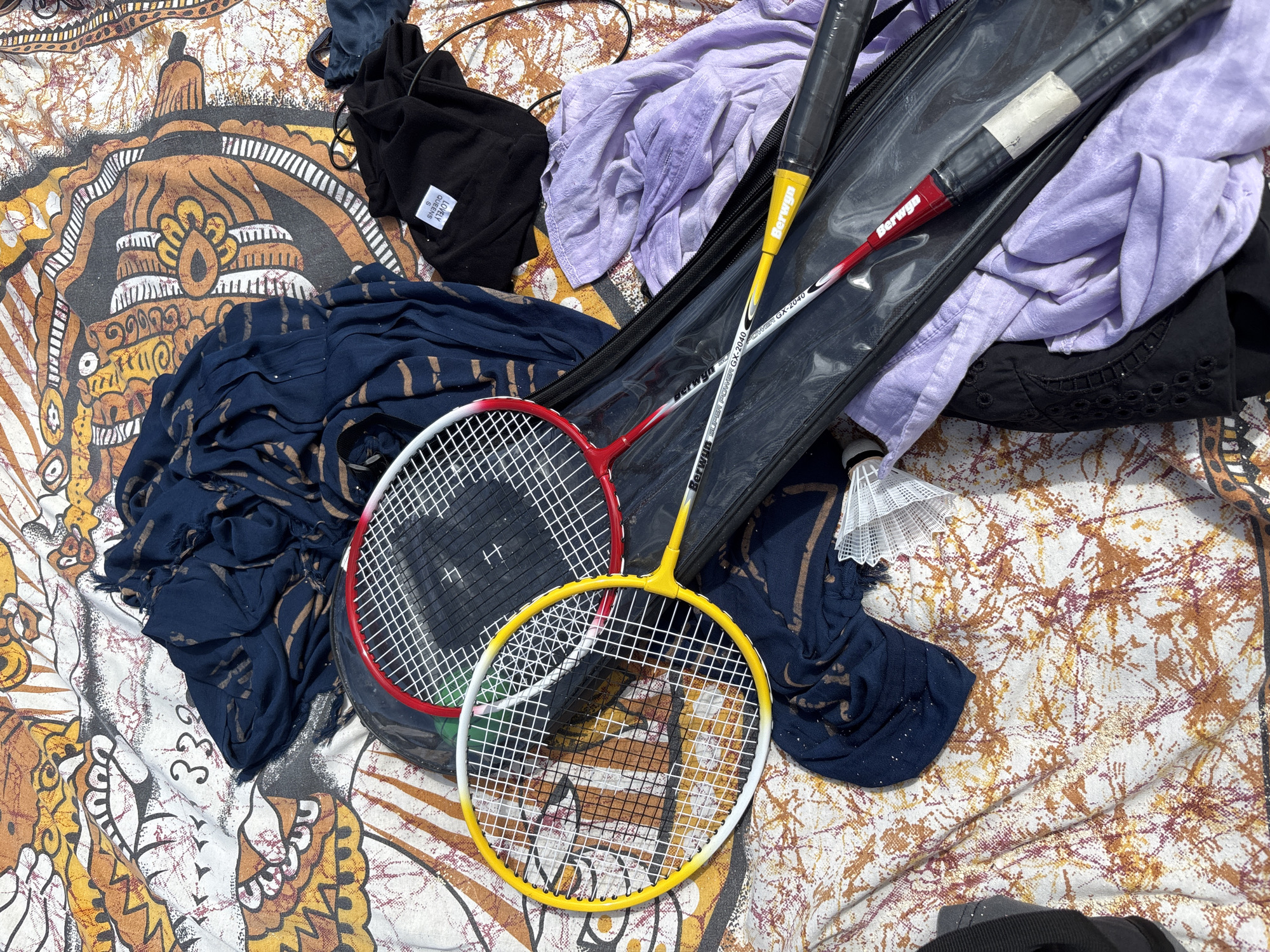 An overhead, medium shot captures a collection of items laid out on a patterned, possibly woven, surface that appears to be a blanket or tapestry. The background features a large, ornate design in shades of orange, yellow, and white, resembling tribal or stylized animal motifs.
Dominating the center of the frame are two badminton rackets, one with a yellow frame and white strings, and the other with a red frame and white strings. Both rackets are lying in a clear plastic badminton racket case. The yellow racket's grip is black and yellow, with the brand name "Berwga" visible. The red racket has a red and black grip, also with "Berwga" printed on it, along with "SUPER POWER GX-2040".
Beside the rackets, a dark blue, loosely folded piece of fabric with a striped pattern is draped. To the right of the blue fabric, a black t-shirt with a white tag that reads "LOVELY QUEENS S" is visible. Further to the right and partially obscuring the rackets, a light purple, wrinkled fabric is laid out. A black garment with eyelet detailing is situated at the bottom right of the frame. A white, lacy edge of another fabric piece is visible on the right side of the image.
The scene is illuminated by bright, natural light, casting shadows that suggest it is daytime. The overall impression is of recreational items and clothing casually placed outdoors. There are no people or animals present in the image.
The location is Kuta Selatan, Indonesia. The scene appears to be an outdoor setting, possibly a beach or a park, given the casual arrangement of items on a patterned surface.