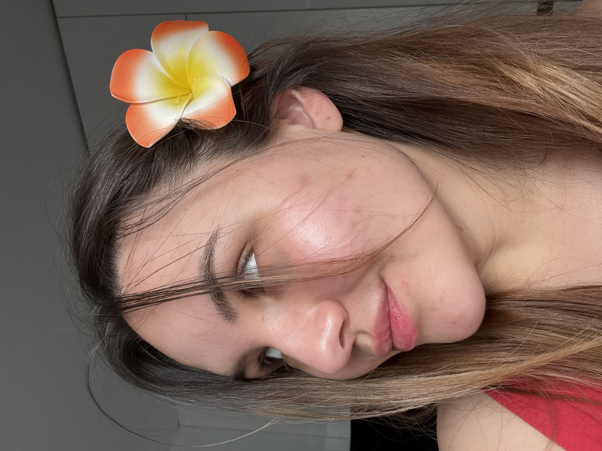 A woman with long, dark brown hair with lighter brown ends is posing for a photo. A flower with orange and yellow petals is attached to her hair near her left ear. She is wearing a red top. The background is a plain gray wall. The image does not contain information that indicates its location.