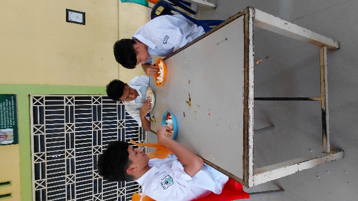 Three young students in white shirts sit around a worn table, eating from bowls. Food spills are visible on the tabletop. Two students are on one side, and the third is on the opposite end. A yellow wall is in the background, adorned with a framed sign and a window with crisscrossing bars. The table is aged, with a white surface and a distressed white frame. A red chair is under one of the students, while the other chairs are not visible.