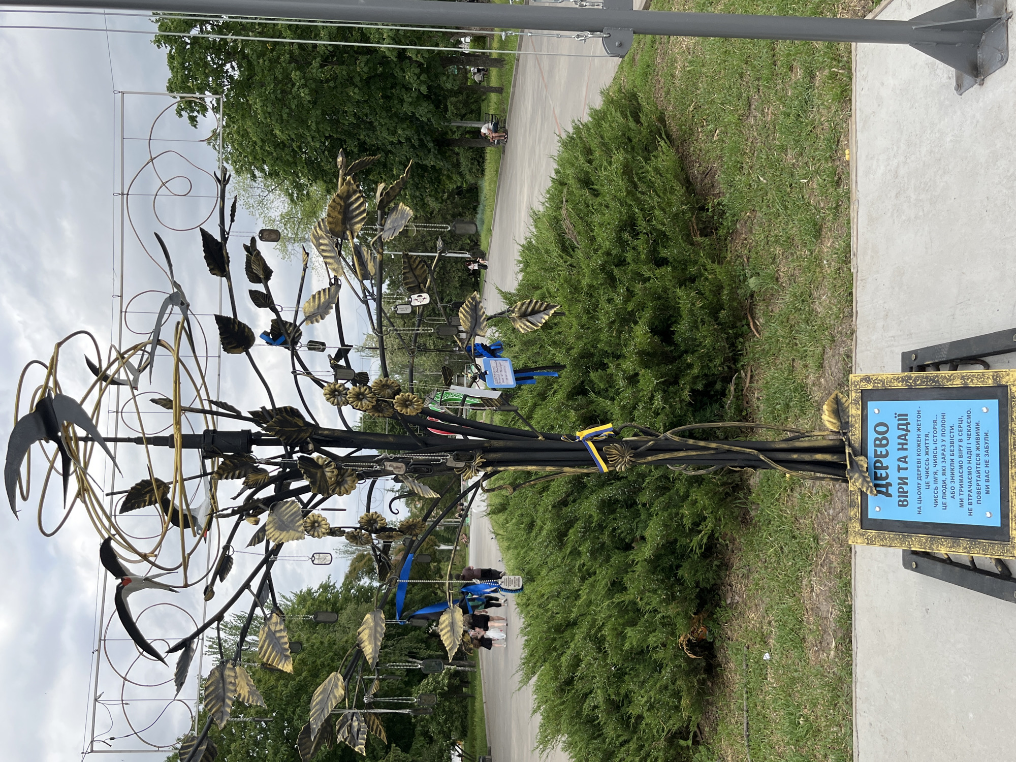 The image shows a metal tree sculpture located on a paved area next to a bush. The tree has metal leaves and decorative elements, including metal birds. Small, rectangular metal tags are hung on the branches. Attached to the tree's base is a framed plaque with text in Ukrainian. The text is legible: "ДЕРЕВО ВІРИ ТА НАДІЇ" (TREE OF FAITH AND HOPE) followed by a message about remembering people in captivity or missing. The background shows a paved path with people walking and trees, suggesting a park or public space. The image shows an object located in the city of Kryvyi Rih.