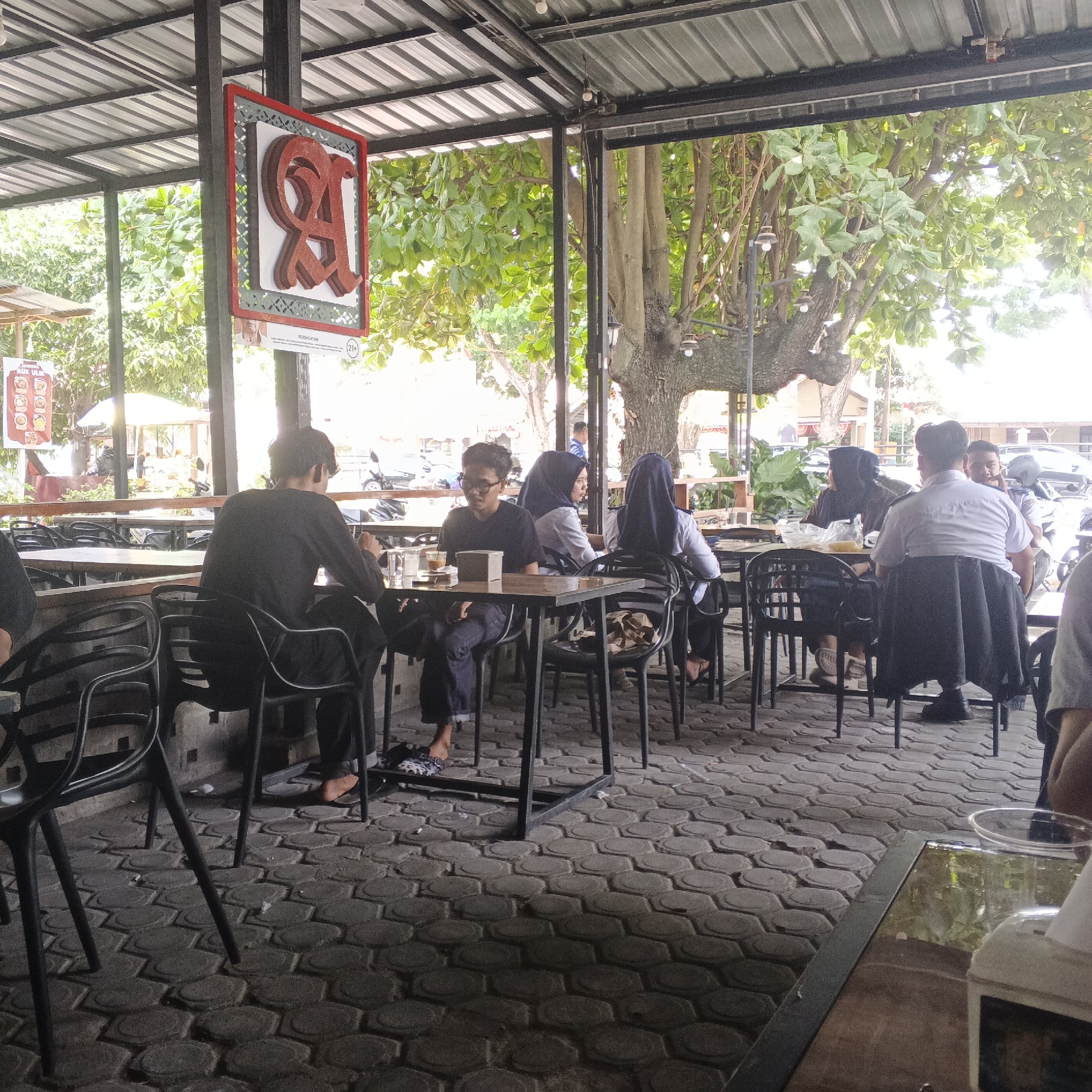 The image depicts an outdoor seating area of a cafe, possibly in Lhokseumawe, Indonesia, judging by the style of the signage and the presence of what appears to be a menu board. The area is covered by a metal roof supported by black metal poles. Sunlight filters through the dense green foliage of trees overhead. The ground is paved with interlocking grey hexagonal bricks.
Several people are seated at dark metal tables, some of whom are dressed in casual attire and headscarves. One individual in the foreground, a person with dark hair wearing a black long-sleeved shirt and dark pants, is seated at a table with a glass and a small box. Another person, also with dark hair, is seated at a table with a white shirt and a black skirt or pants. To their right, a person wearing a white shirt and a dark headscarf is looking away. Further back, more patrons are visible, with one person wearing a white collared shirt and dark pants. Various tables and black metal chairs are arranged throughout the seating area. A sign with a stylized red "RA" logo and decorative border hangs from a pole. In the background, a menu board displaying food items and a "21+" warning is visible. The overall atmosphere suggests a casual dining or gathering place.