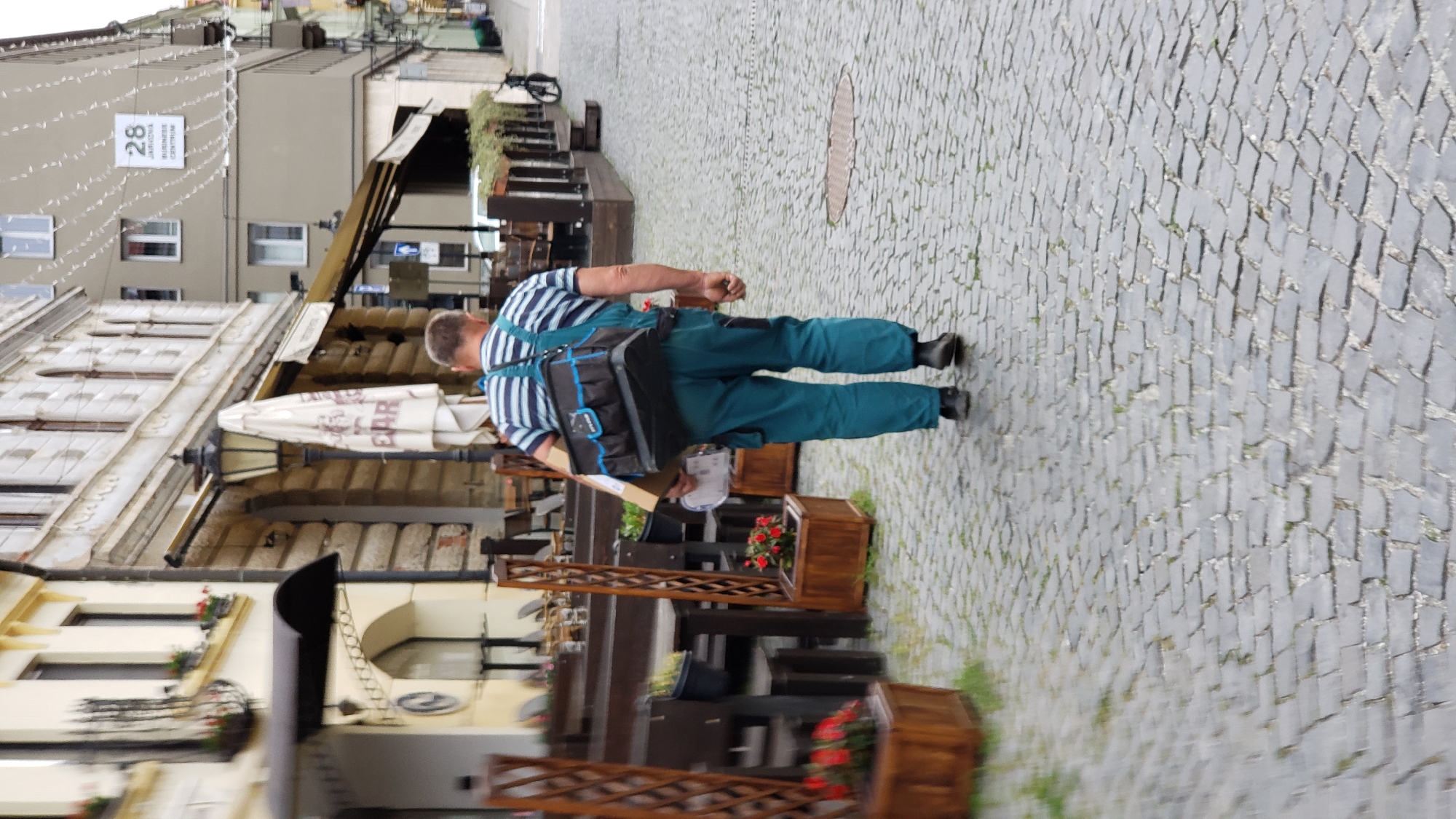 A person wearing blue overalls and a striped shirt walks down a cobblestone street in Prešov, Slovakia. The individual carries a large black and blue toolbox on their left shoulder and a cardboard box in their right hand. The street is lined with buildings featuring architectural details such as arches and ornate facades. Outdoor seating areas with tables and chairs belonging to restaurants are visible on the left side of the street. In the background, a modern building with signage displaying "28" is present. The weather appears overcast.
