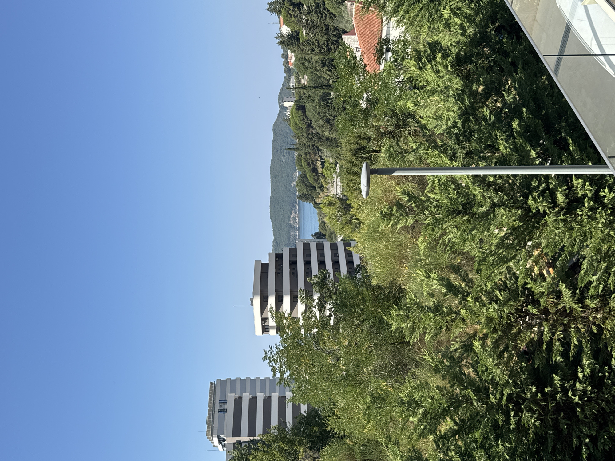 This is a landscape photograph taken in Boreti, Montenegro, on a clear, sunny day. The scene is dominated by lush green foliage in the foreground, with several trees and bushes obscuring the lower half of the image. In the mid-ground, two modern multi-story buildings stand out. The taller building on the left has a dark gray and white striped facade with visible solar panels on its roof. The shorter, wider building to its right also features a similar striped design. Beyond the buildings, a body of water, likely the Adriatic Sea, is visible, with a distant green, mountainous island or peninsula in the background. The sky is a bright, cloudless blue. A modern grey lamppost is positioned in the foreground, and a glass balcony railing is visible in the bottom right corner, suggesting the photo was taken from an elevated vantage point. The overall impression is one of a coastal urban environment surrounded by nature. There are no people visible in the photograph.