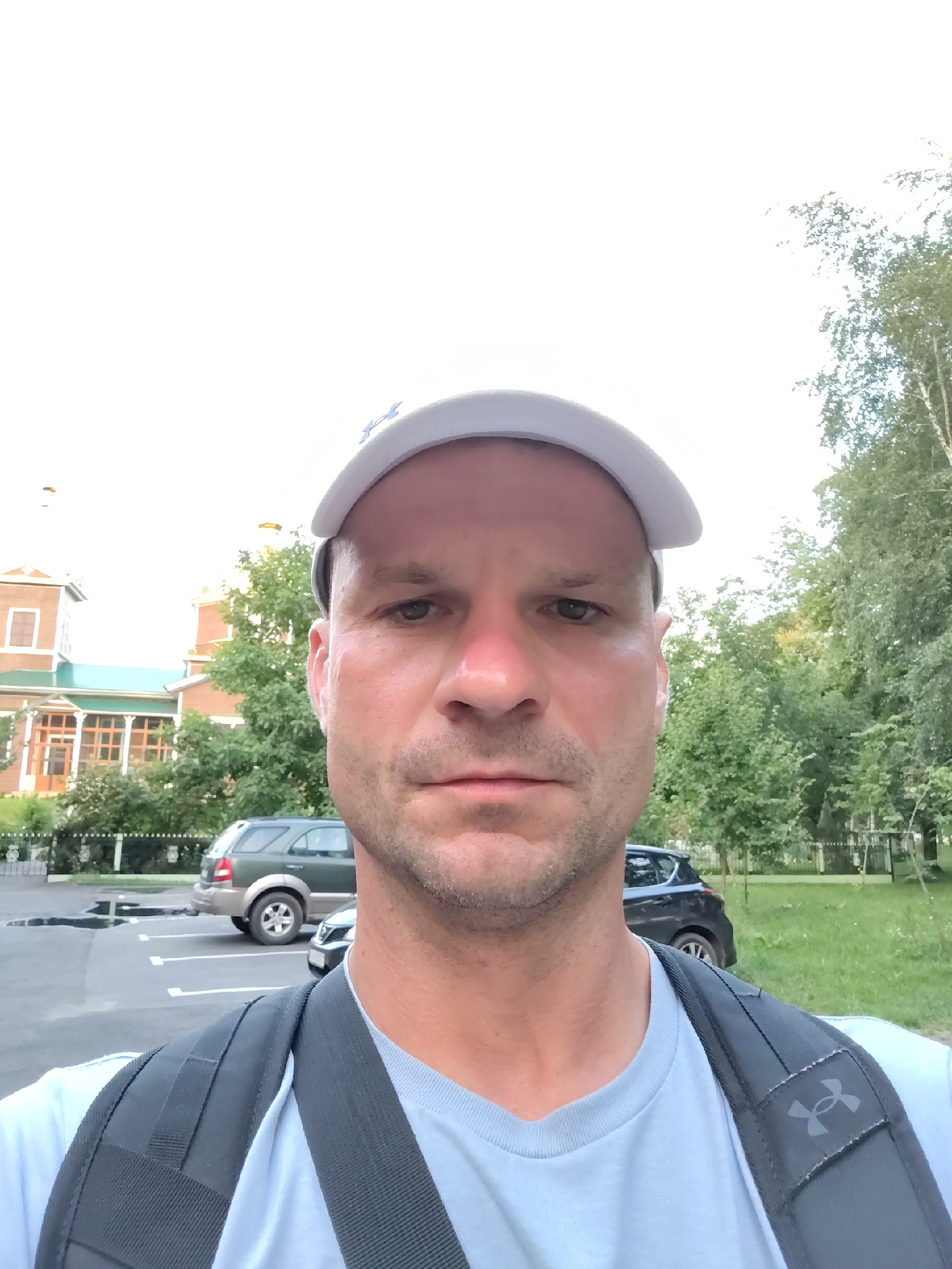 A man wearing a light blue t-shirt and a white baseball cap is taking a selfie outdoors. He has short brown hair and is looking directly at the camera. In the background, there is a two-story building with a green roof, trees, a parking lot with two cars, and a grassy area. The scene appears to be a park or residential area. The man is wearing a backpack with the Under Armour logo visible.