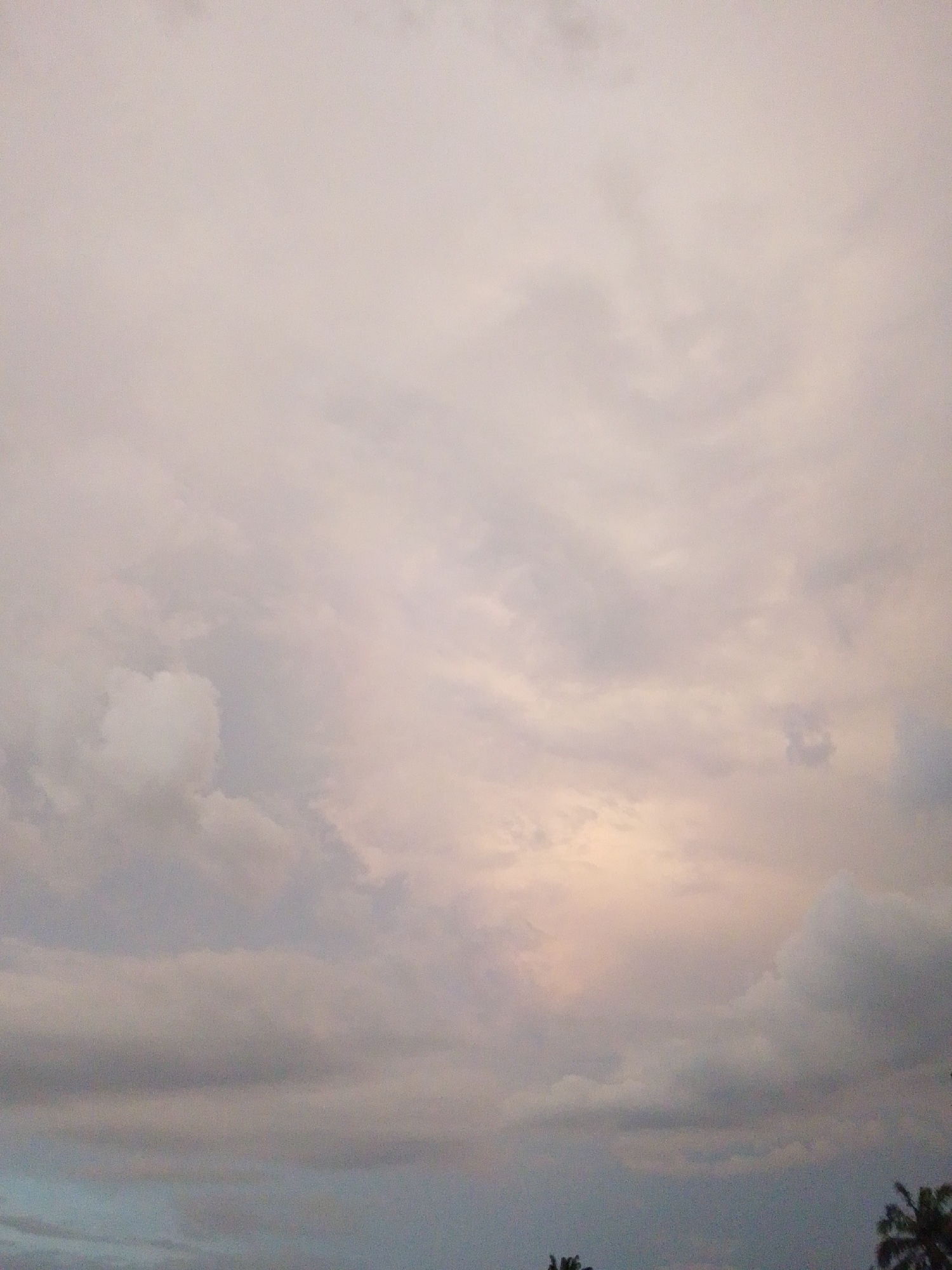The image shows a cloudy sky over Serti, Nigeria. The clouds are a mix of white, gray, and pink hues, suggesting either sunrise or sunset. The sky occupies the majority of the frame, with the tops of some palm trees visible at the very bottom edge of the image. There are no people or identifiable objects in the scene. The overall impression is one of a natural landscape in the early morning or late evening.