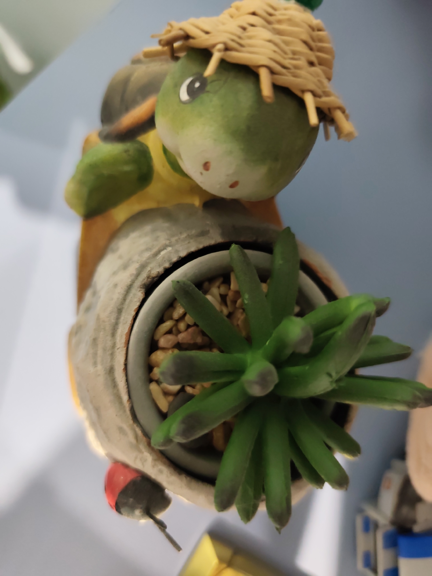 A close-up, top-down shot captures a decorative ceramic figurine, resembling a green-headed creature, possibly a turtle or frog, wearing a light beige, woven straw hat. The creature's head is predominantly green with a prominent white and black eye, and small reddish-brown speckles on its lower face. It appears to be 'carrying' a distressed grey, concrete-textured pot, which contains numerous small, light brown pebbles and a vibrant green artificial succulent plant with multiple fleshy leaves, some displaying darker gray tips. A small red and black decorative ladybug is situated on the lower left side, adjacent to the pot. The background is blurred, showing soft blue tones in the upper left and indistinct lighter shapes on the right. This indoor scene is well-lit, highlighting the textures and colors of the figurine and plant. This scene is located in Pazardzhik, Bulgaria.