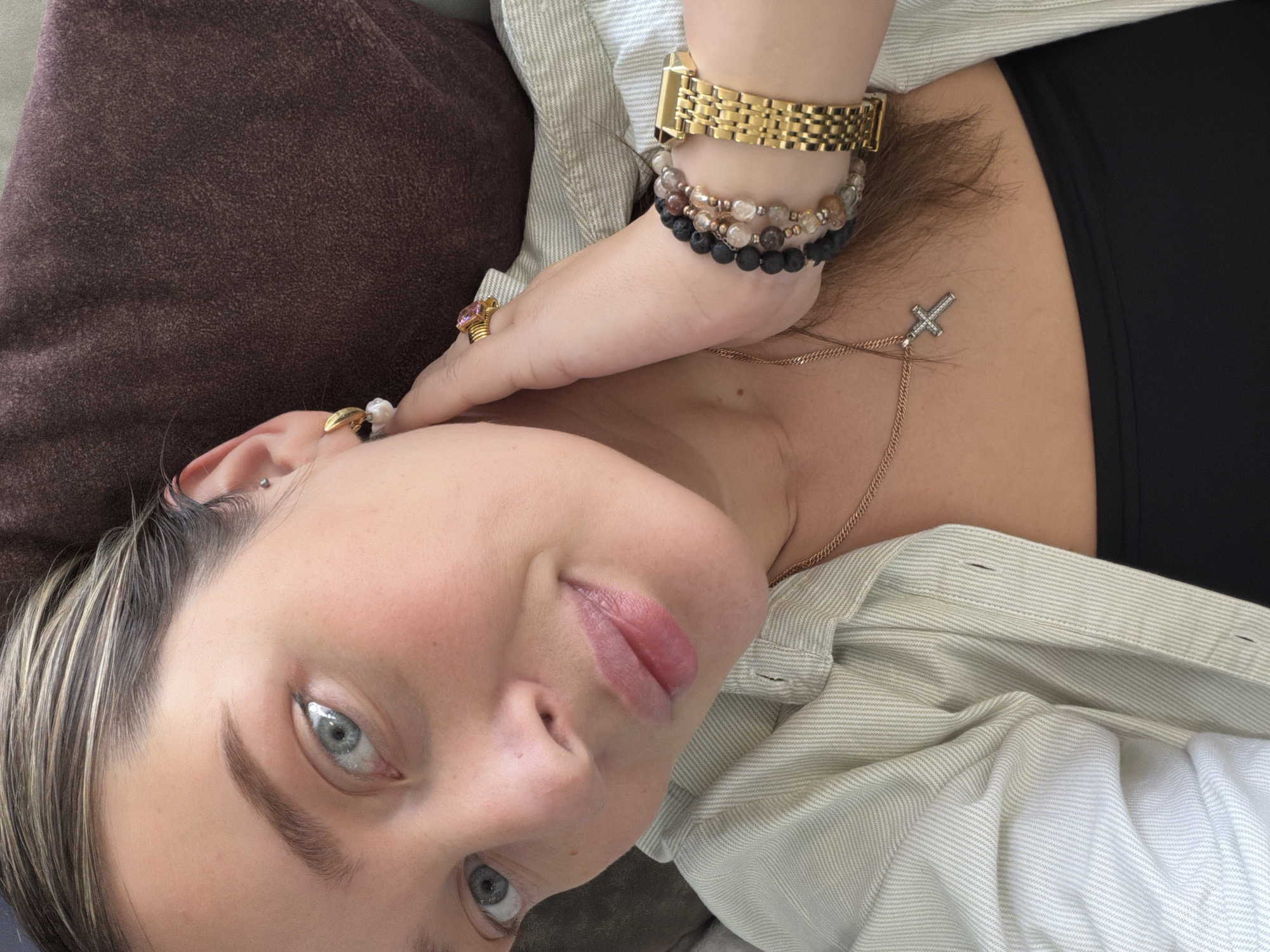 A selfie-style, close-up image features a fair-skinned individual with light hair and blue eyes. They are lying down, looking at the camera with a slight smile. They wear multiple bracelets on one wrist, including a gold watch, along with rings on their fingers. A delicate gold necklace with a cross pendant rests on their chest. The person wears a light-colored, striped button-down shirt, and a dark-colored top is visible underneath. A brown cushion is in the background. There is no evidence in the photo to suggest a location in the city of Chaikovsky, Russia.