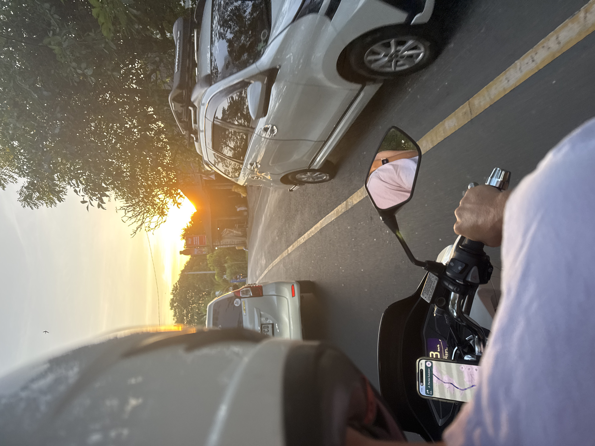 A view from a motorcycle at sunset, showing a road with a white car to the right and another car in front, farther away. The motorcycle rider is holding the handlebar, and a smartphone is mounted on the motorcycle, displaying a map of "Jl. Pantai Suluban." The motorcycle's rearview mirror reflects the rider's light pink shirt. The sun is setting ahead, between trees.