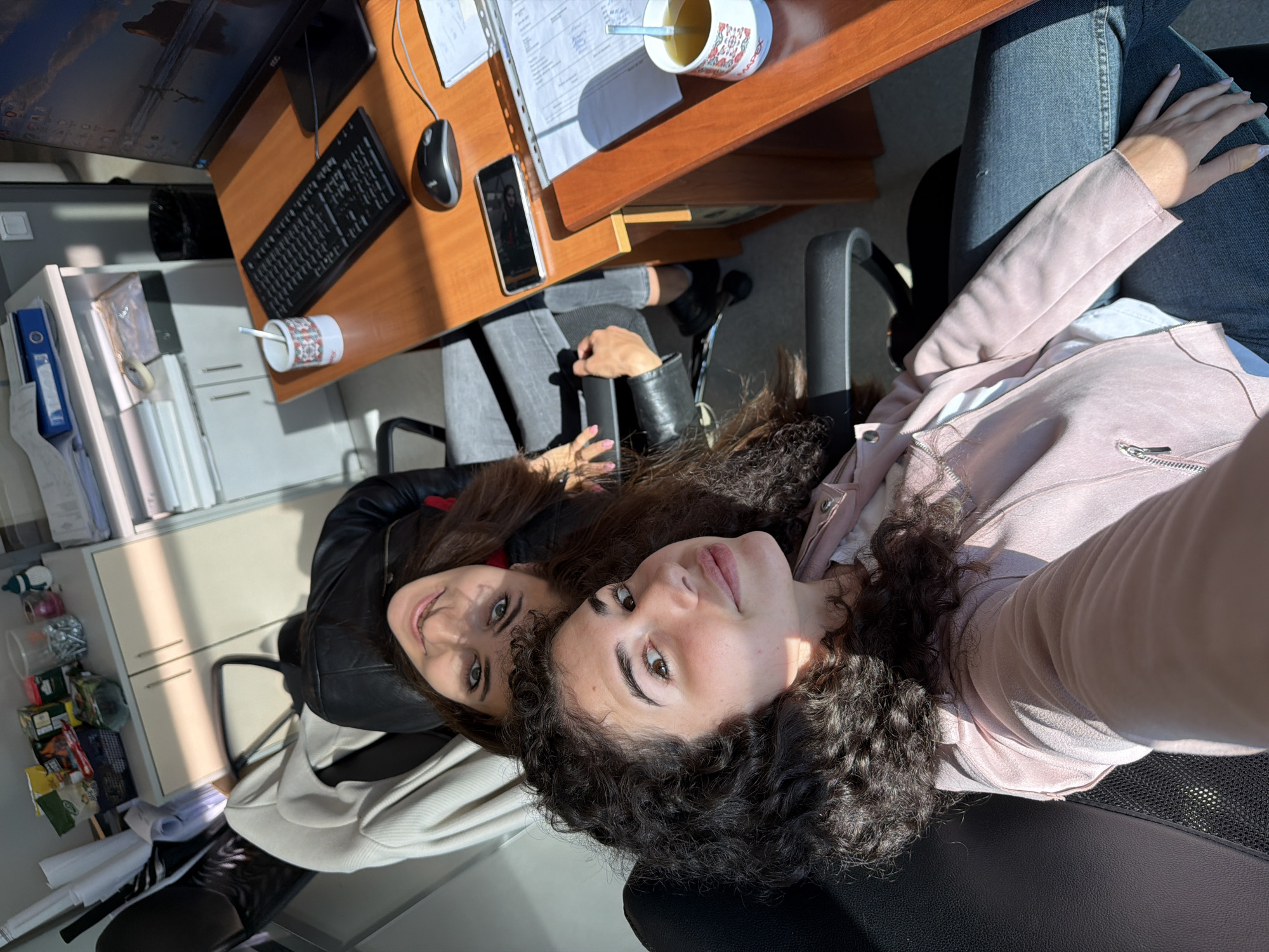 Two young women are captured in a selfie taken in an office environment that appears to be in Sofia, Bulgaria. The woman in the foreground, with curly dark hair and wearing a light pink jacket over a white shirt and blue jeans, is looking directly at the camera with a slight smile. Behind her, slightly out of focus, is another woman with long dark hair, wearing a black leather jacket and jeans, also looking towards the camera.
They are seated in office chairs, suggesting they are colleagues or friends taking a break. The background reveals a typical office setting with desks, a computer monitor displaying a nature scene, a keyboard, a mouse, a mobile phone, stacks of papers, and office supplies on shelves. Sunlight streams into the room, casting long shadows and highlighting dust particles in the air. A mug of tea with a spoon is visible on the desk. The overall impression is one of a casual moment captured during a workday.