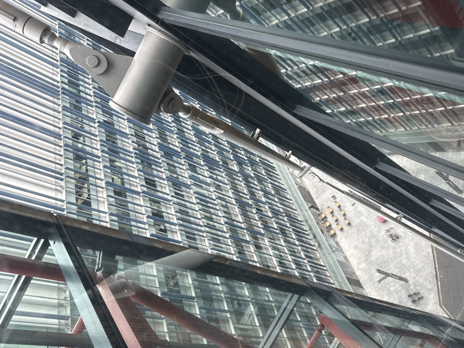 The image presents an oblique, high-angle view looking down into an urban plaza from within a modern high-rise building. In the immediate foreground, large glass panels display reflections of architectural features, including diagonal red structural beams and a prominent grey steel tension rod with articulated bolted connections extending downwards. Across the plaza, a multi-story building facade is visible, characterized by numerous closely spaced, vertical, light-colored fins or louvers that span its height, partially obscuring the windows of internal office spaces. The ground-level plaza is paved with light grey material and contains several clusters of outdoor tables with yellow/orange chairs. Multiple bicycles are parked at two distinct locations within the plaza, one group adjacent to a red utility box. Two indistinct human figures are discernible walking on the far left side of the plaza. A metallic 'Y'-shaped structure is also present on the paving. This scene is located at The Leadenhall Building, 122 Leadenhall Street, City of London, United Kingdom.