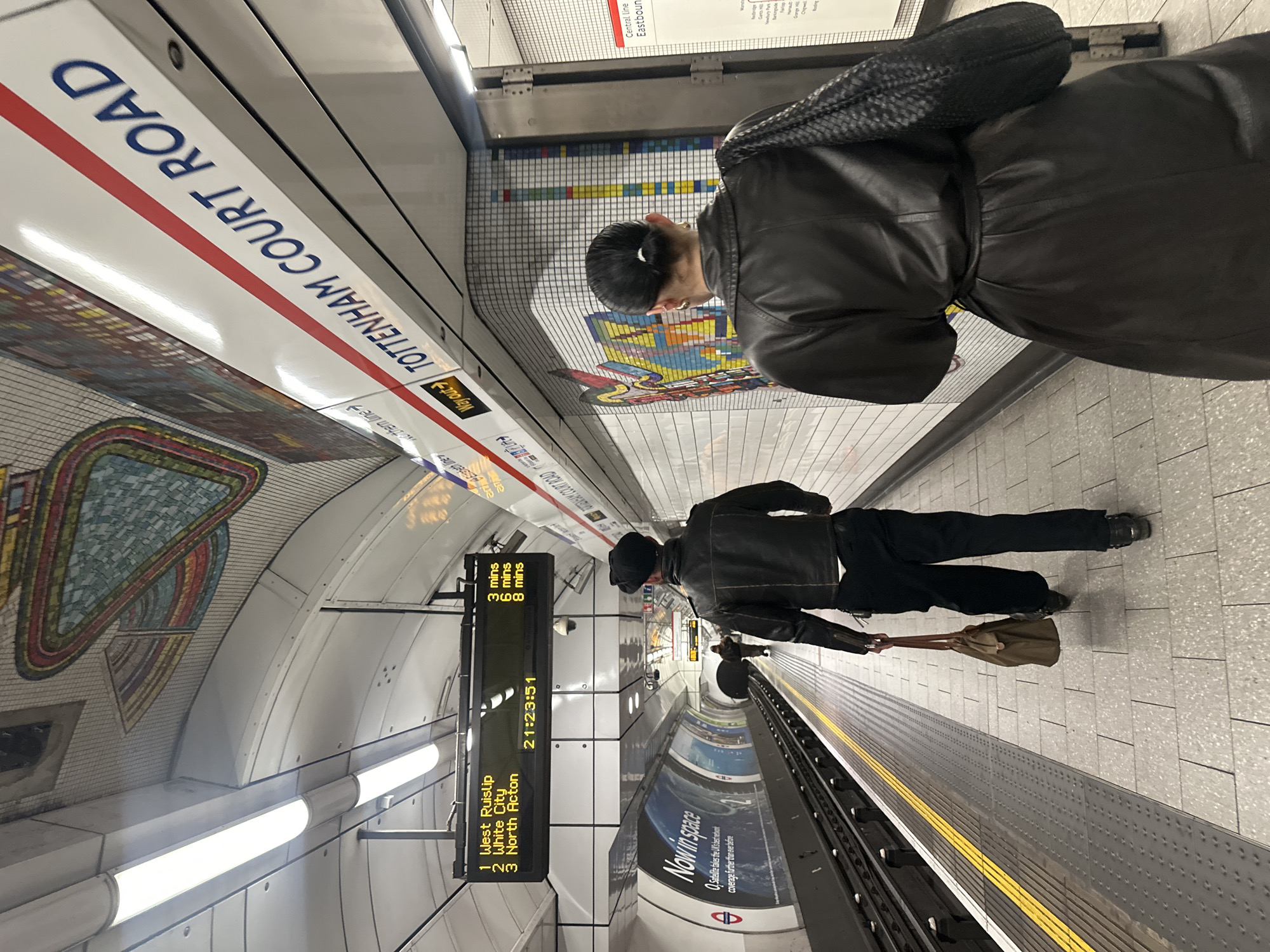 The image shows the interior of a London Underground station in the United Kingdom, specifically Tottenham Court Road station. A sign identifies the station's name in bold, blue letters. There are passengers on the platform; one wearing a black leather coat. A digital display shows upcoming train destinations: West Ruislip, White City, and North Acton, along with their estimated arrival times and the current time of 21:23:51. An advertisement for coverage further than ever before is displayed on the platform wall. Yellow stripes are shown near the platform edge as a safety precaution.