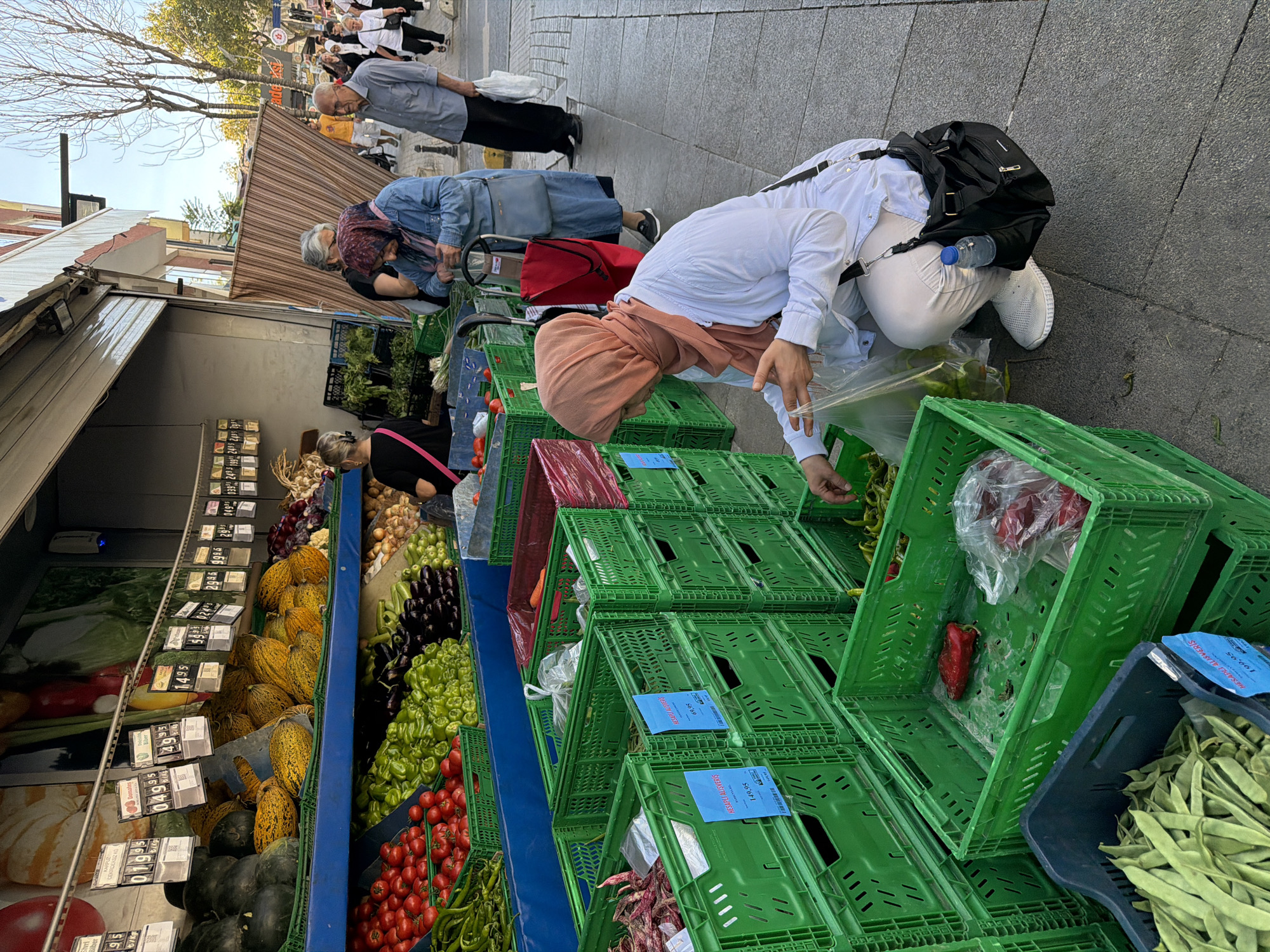 An outdoor market scene in Kadıköy, Turkey, features a variety of fresh produce displayed in green plastic crates. In the foreground, a woman wearing a white lab coat and a light orange headscarf is squatting, selecting green peppers from a crate. She holds a clear plastic bag in her right hand. A black messenger bag with a water bottle attached hangs from her shoulder. Behind her, stacked green and red crates hold tomatoes, peppers, and other vegetables. Further back, a display of melons and watermelons is visible, along with onions and other produce. Several shoppers are present, including a woman in a denim jacket and headscarf with a shopping cart, and an elderly man in a light blue shirt and dark trousers. The market appears to be set up along a paved sidewalk next to a building with an open storefront. The overall lighting suggests it is daytime, with sunlight casting shadows.