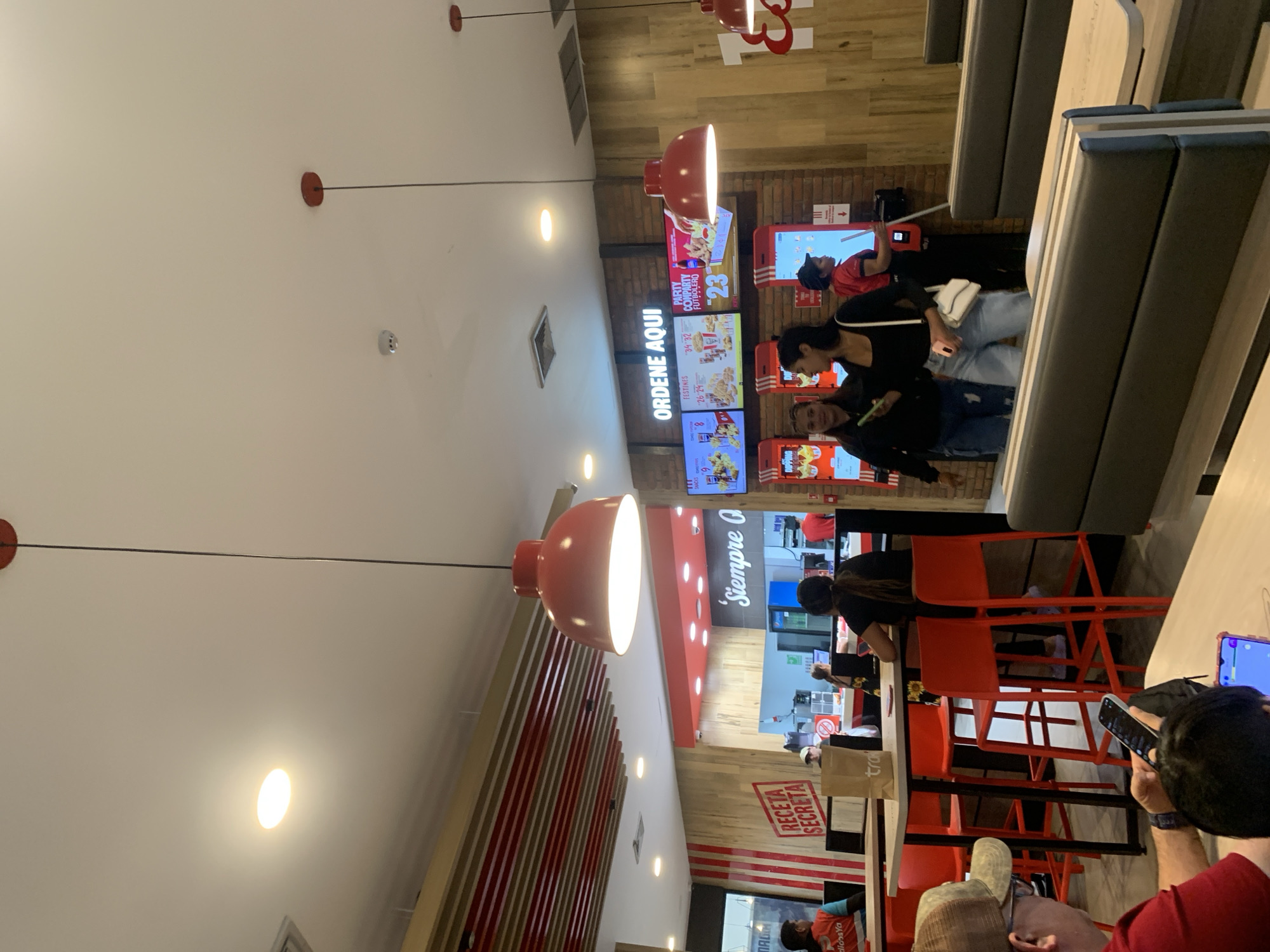 This image depicts the interior of a fast-food restaurant, likely a KFC, in Porlamar, Venezuela. The scene is brightly lit with overhead fluorescent lights and large red pendant lamps. In the foreground, a man wearing a red shirt and a camouflage cap is seated at a table, looking at his smartphone. Another person, partially visible, is seated next to him, also looking at a phone. To the right, two young women are standing near a digital menu board. One is wearing a black top and light blue jeans, carrying a white purse. The other is in a black hoodie and jeans. Behind them, a staff member in a red uniform stands near a self-ordering kiosk.
The restaurant features red and white branding, with wooden accents and brick walls. Digital menu boards display food items and prices. In the background, a service counter with a blue refrigerator is visible, and other patrons can be seen. High-top tables with orange stools are set up in the dining area. The overall environment suggests a busy, modern fast-food establishment.